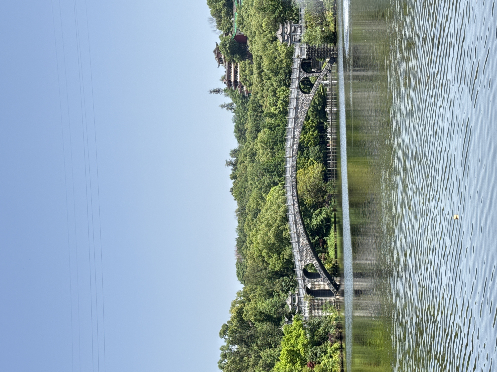 The image captures a serene landscape featuring a prominent arched bridge crossing a body of water. The bridge is constructed of stone with distinctive circular openings along its span. Dense green foliage covers the land on the bridge's far side, with building structures visible among the trees, including traditional Chinese architecture. The water's surface reflects the sky and surrounding greenery, exhibiting gentle ripples. Electrical lines can be seen stretching across the clear blue sky, and the landscape is located in Jiulongpo District, China.