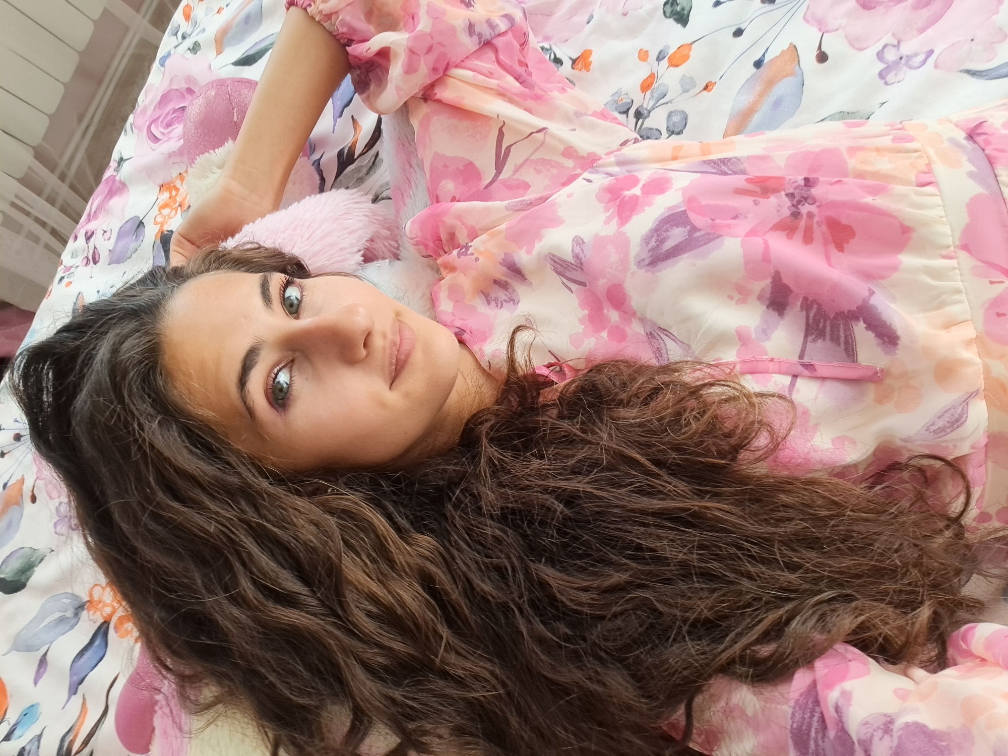 A young woman with long, dark, curly hair lies on her back on a bed with a floral comforter. She is wearing a light pink dress with a floral pattern. Her head is tilted to the side, and her eyes are open and looking at the camera. She has a slight smile on her face. Her right arm is bent at the elbow, and her hand is resting on her head. The background is blurred, but it appears to be a bedroom. The lighting is soft and natural, suggesting it could be daytime. The overall mood is relaxed and comfortable.
