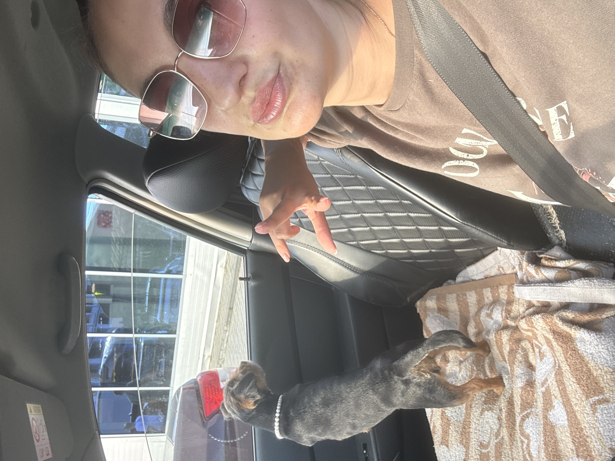 A woman in sunglasses and a brown sweatshirt is taking a selfie inside a car. Next to her, a small black dog with a pearl necklace is standing on the car seat, looking out the window. The car is parked, and through the windows, a building with large glass panes reflecting the sky can be seen. The scene appears to be in an urban environment, possibly a parking lot.