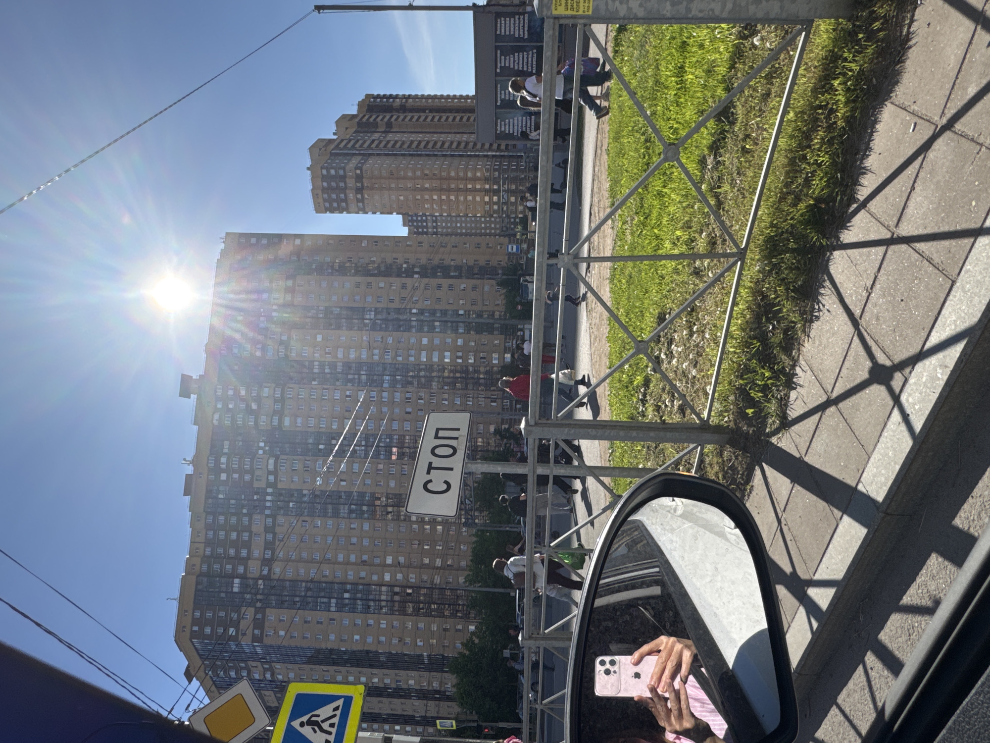 The image captures an urban street scene on a bright, sunny day, viewed from inside a vehicle. In the foreground, a car door frame and a side mirror are visible. The mirror reflects a person's hands holding a pink smartphone (an iPhone with three rear cameras) and part of a striped garment.
Beyond the vehicle, a metal fence with a diamond-grid pattern separates a strip of green grass from a paved sidewalk. The fence casts prominent, geometric shadows across the sidewalk, indicating a high sun position. Several people are visible on the sidewalk, walking or standing, some partially obscured by the fence or shadows.
In the mid-ground, a white rectangular sign with black Cyrillic letters reads "СТОП" (STOP), mounted on a pole near the fence. Further to the bottom left, two road signs are partially visible: a yellow diamond shape (indicating a priority road) and a blue square with a white pedestrian symbol (for a crosswalk).
The background is dominated by several tall, multi-story residential buildings, appearing in light brown and darker tones, with numerous windows. The bright sun is prominently featured in the upper left quadrant of the sky, creating a strong lens flare. The sky is clear and blue. The Cyrillic text on the "СТОП" sign points to a Russian-speaking region, consistent with the provided context of Saint Petersburg, Russia.