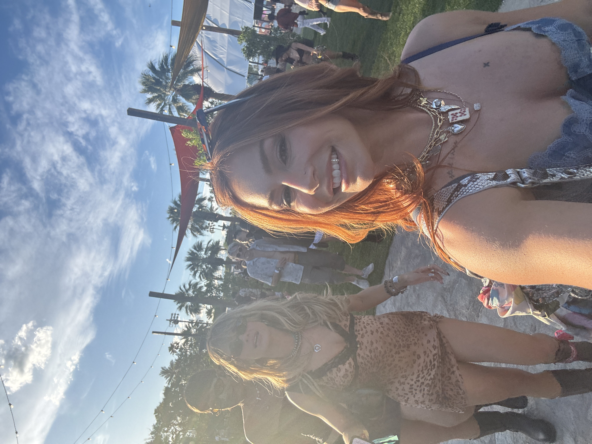 The image captures a group of people outdoors in La Quinta, United States of America. A woman with auburn hair is in the foreground, smiling and facing the camera. She is wearing a necklace and a top with lace detailing. Behind her, another woman with blonde hair is wearing a leopard print dress and sunglasses. In the background, there are several other individuals, palm trees, and a tent-like structure. String lights are visible overhead, and the sky is blue with scattered clouds.