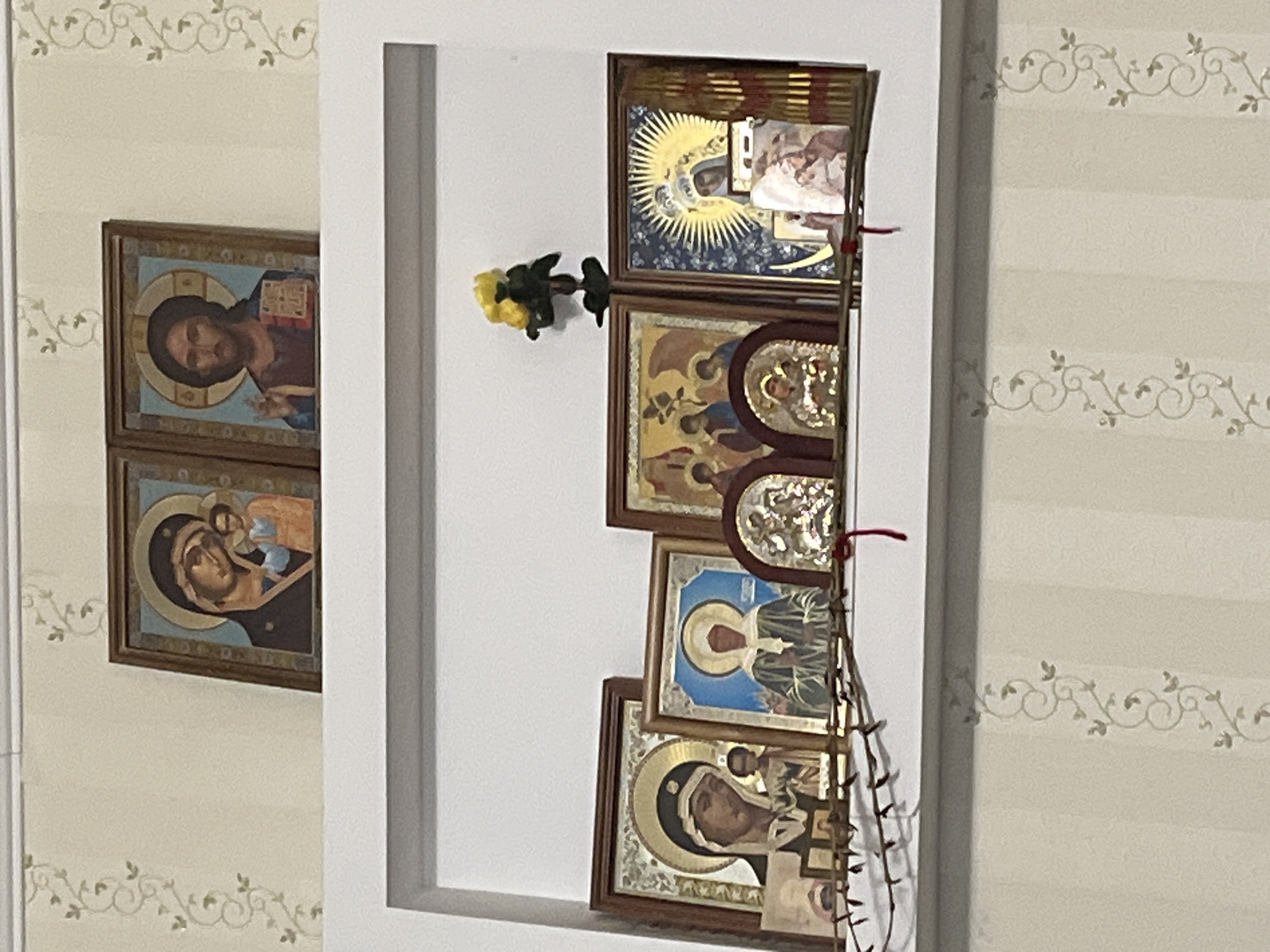 The image shows a shelf with several religious icons. On the top shelf, there are two framed icons, one depicting the Virgin Mary and Child, and the other depicting Jesus Christ. On the lower shelf, there are several more icons, including one of the Virgin Mary and Child, another of a saint in a white veil, and two smaller golden icons depicting religious scenes. There is also a yellow artificial flower on the right side of the lower shelf. The shelf is mounted on a wall with a light-colored wallpaper featuring a subtle vine pattern. The scene appears to be an indoor home altar or devotional space. The image is a photograph taken from a slightly upward angle.