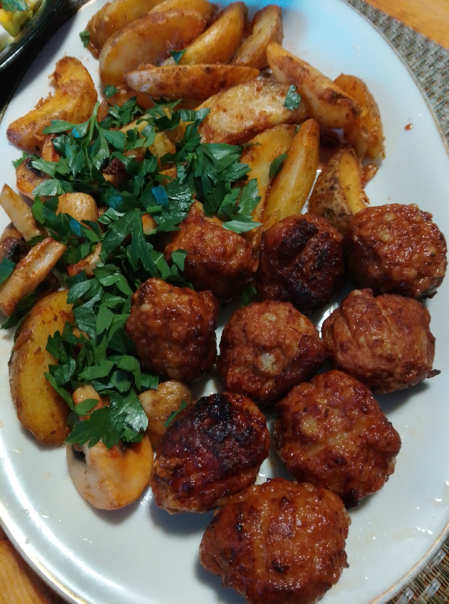 A white oval plate holds a meal consisting of roasted potato wedges, meatballs, and chopped parsley. The potato wedges are golden brown and seasoned, scattered around the upper portion of the plate. Below the potatoes are several round, browned meatballs, some showing a glistening sauce. A generous amount of fresh, chopped parsley is sprinkled over the potatoes and some of the meatballs. A few sliced mushrooms are also visible among the potatoes. The plate is set on a textured placemat, and the scene appears to be indoors, possibly in a dining setting in the city of Wedel, Germany.
