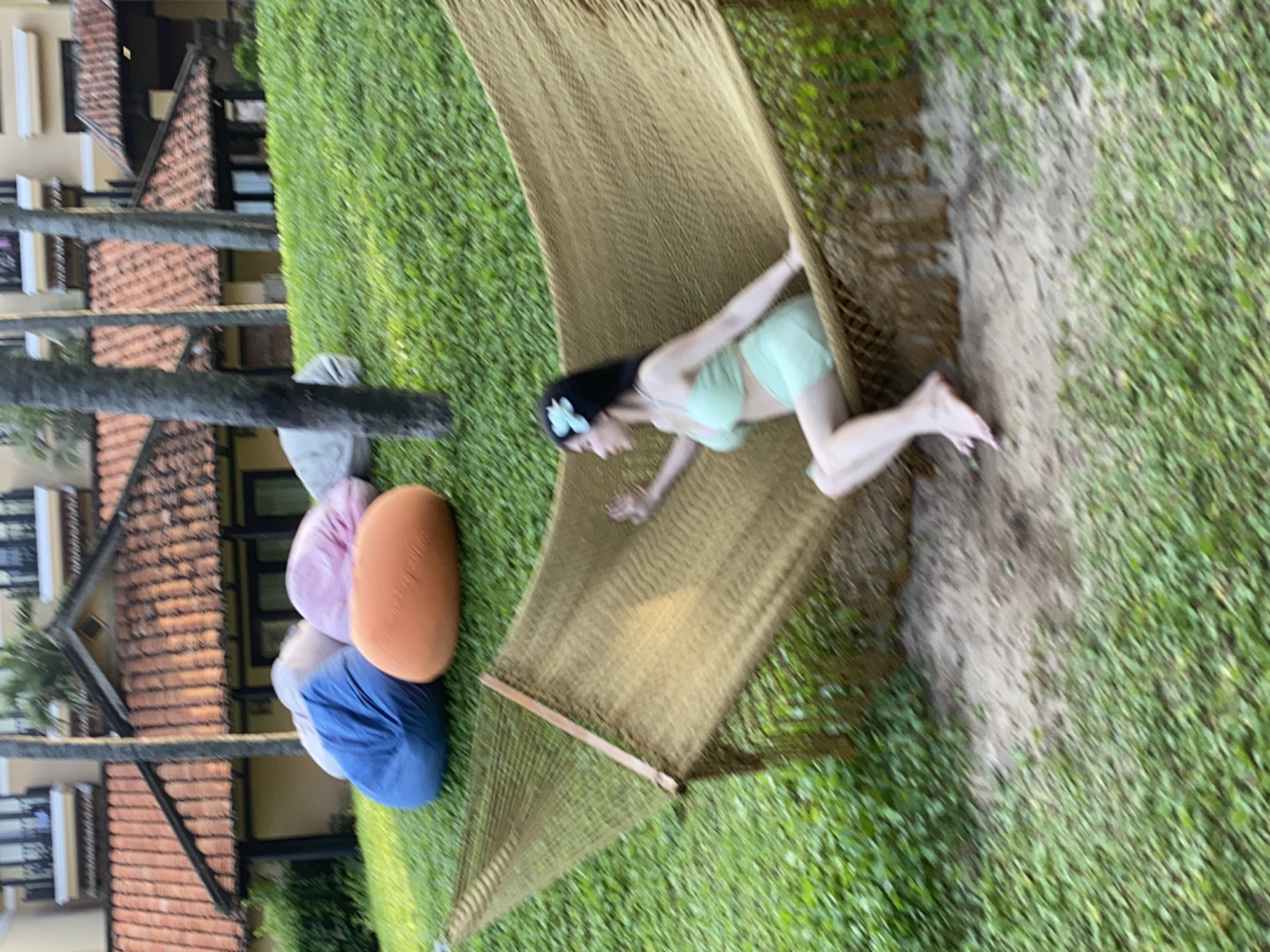 The image depicts an outdoor leisure area in Phu Thuy Ward, Vietnam. In the lower right foreground, a woman with dark hair, adorned with a light blue flower clip, is seated in a large, dark brown woven hammock. She wears a light green two-piece outfit consisting of a bra top and shorts. Her bare feet are visible, resting on the sandy ground beneath the hammock. Her right arm is slightly raised, and she faces towards the left side of the frame. The hammock is situated over a patch of light brown sand and dirt, which is surrounded by green grass. Behind the hammock, a grassy slope rises, featuring several large beanbag chairs in various colors, including prominent blue, orange, and pink ones. In the background, behind the grassy incline, multiple multi-story buildings with reddish-brown tiled roofs are visible, interspersed with tall palm trees. The scene is well-lit, indicating daytime.