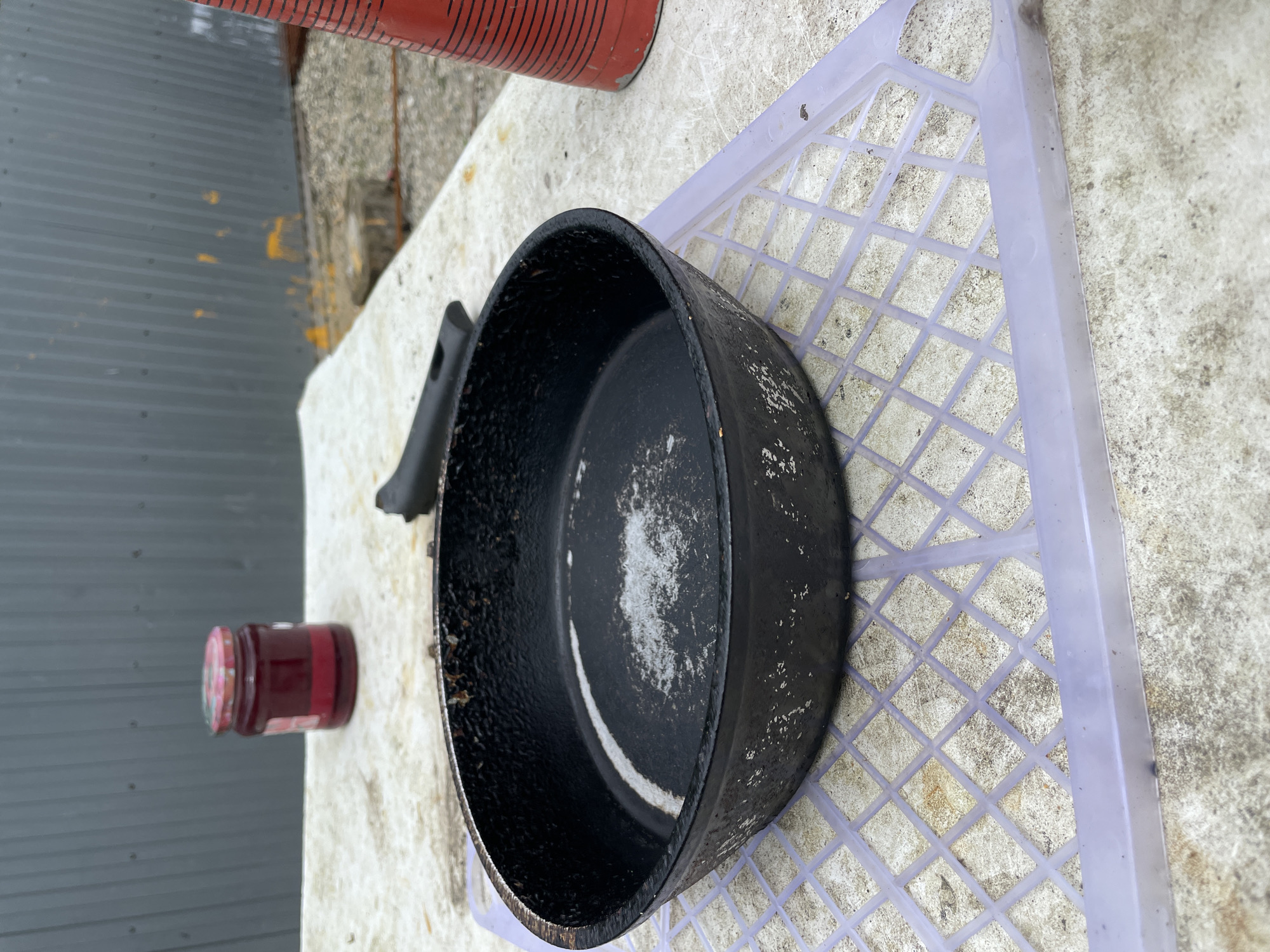 Olginka, Russia. A black frying pan sits on a white, lattice-patterned plastic tray atop a white, possibly concrete surface. A jar of red jam is adjacent to the pan, and a red and black striped container is partially visible in the upper left corner. The background includes a gray, corrugated metal wall. The lighting suggests an outdoor setting.