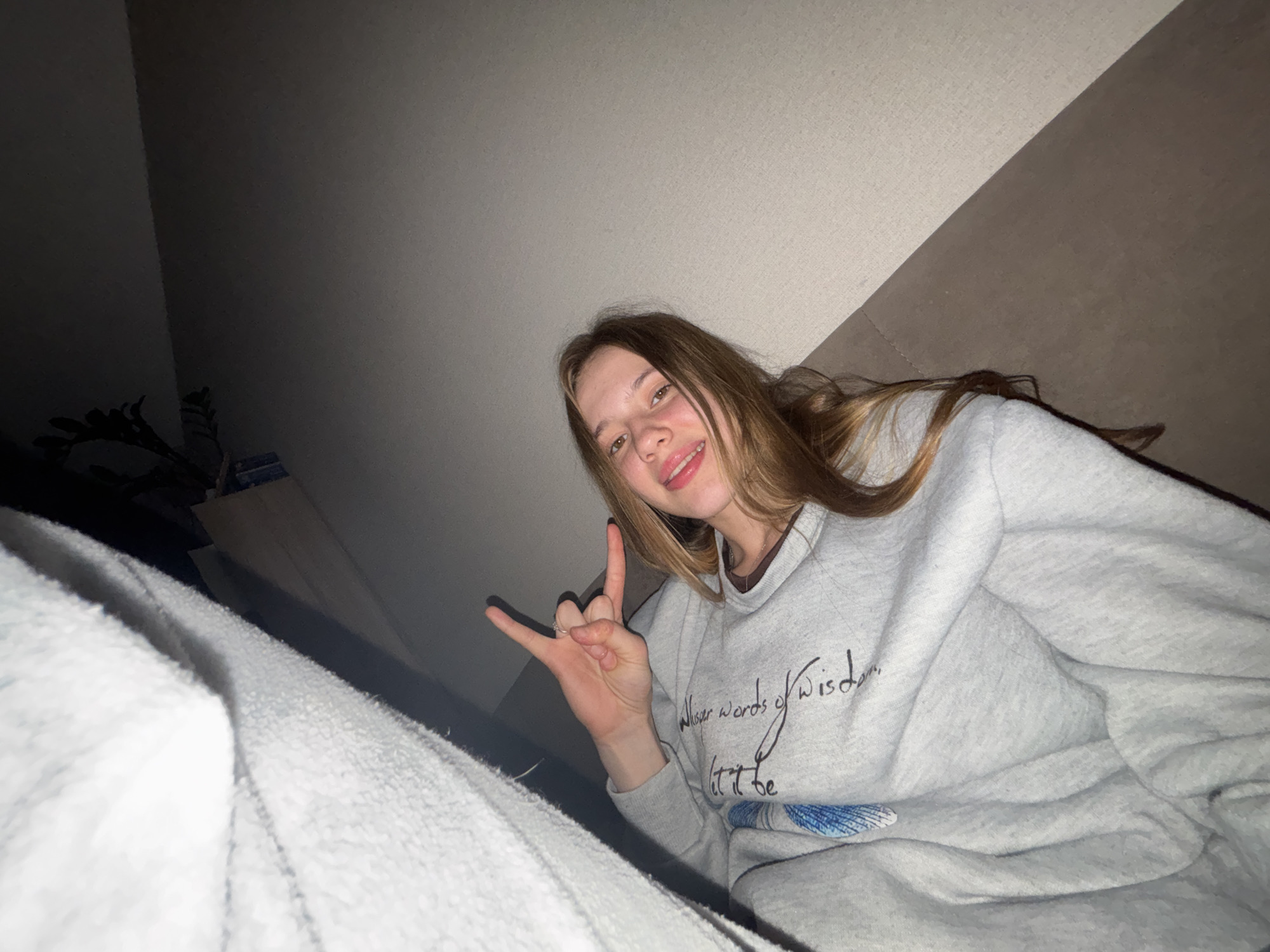 A young woman is pictured from a low angle, lying on a couch in what appears to be a bedroom. She is wearing a light gray sweatshirt with a dark brown collar. Her right hand is raised, forming a "rock on" gesture with her index and pinky fingers extended, while her middle and ring fingers are curled down and her thumb is extended. She has long, wavy brown hair and is smiling at the camera. Her left arm is tucked underneath her. The background consists of a wall and a couch. A white blanket or sheet is draped over the left side of the frame, partially obscuring the wall. The lighting is dim, suggesting it may be nighttime or the room is not well-lit. There are no visible indicators to determine the specific city or country.