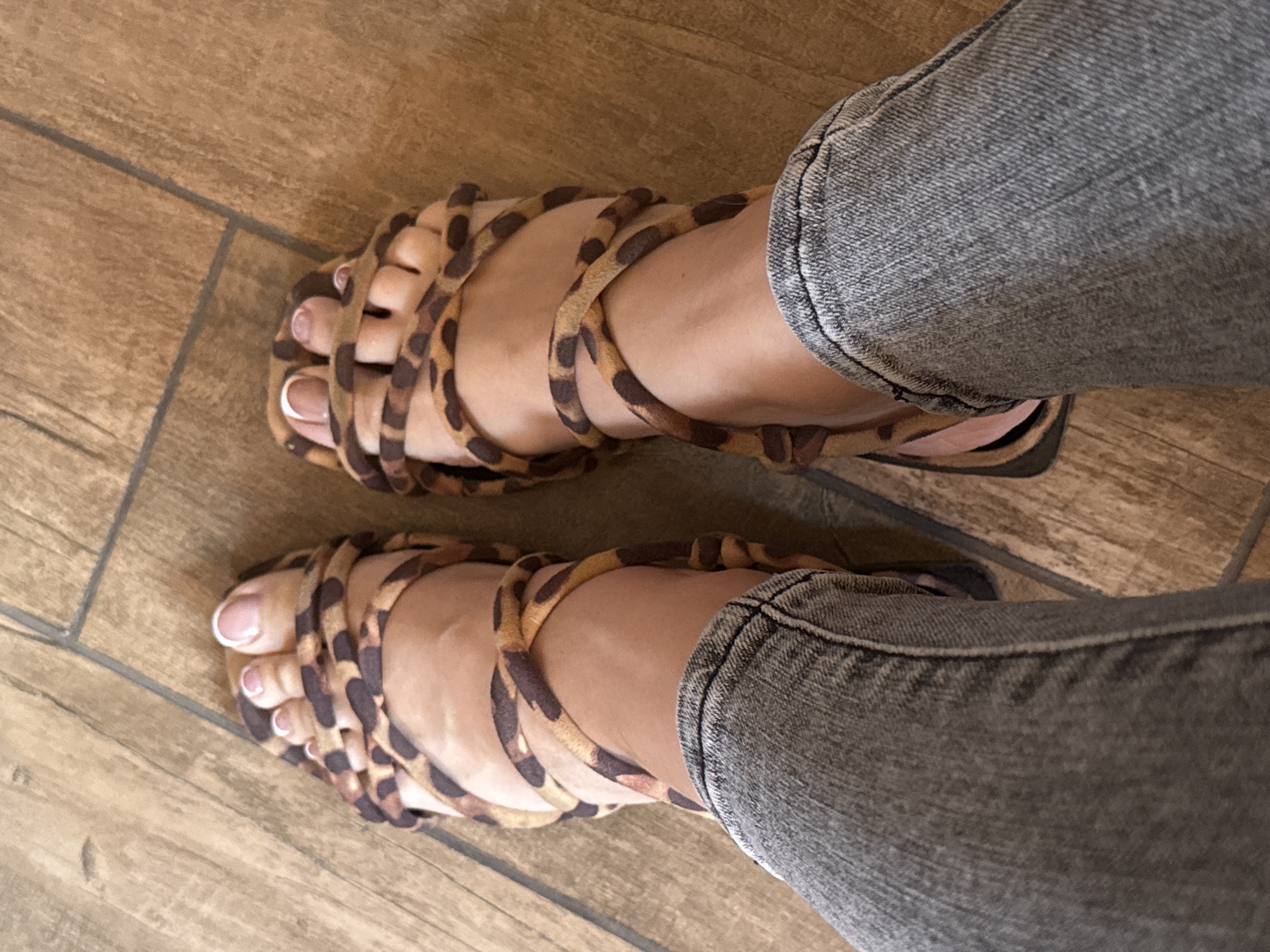 A close-up, overhead view shows a pair of feet adorned with leopard-print sandals and French-manicured toenails, resting on a light brown wooden floor. The feet are clad in gray denim jeans that end just above the ankles. The perspective suggests the person is sitting or has their legs extended. The scene is indoors, with the lighting appearing to be natural or from an overhead source, casting minimal shadows. There are no other individuals or objects visible in the frame, nor any identifiable urban landmarks.