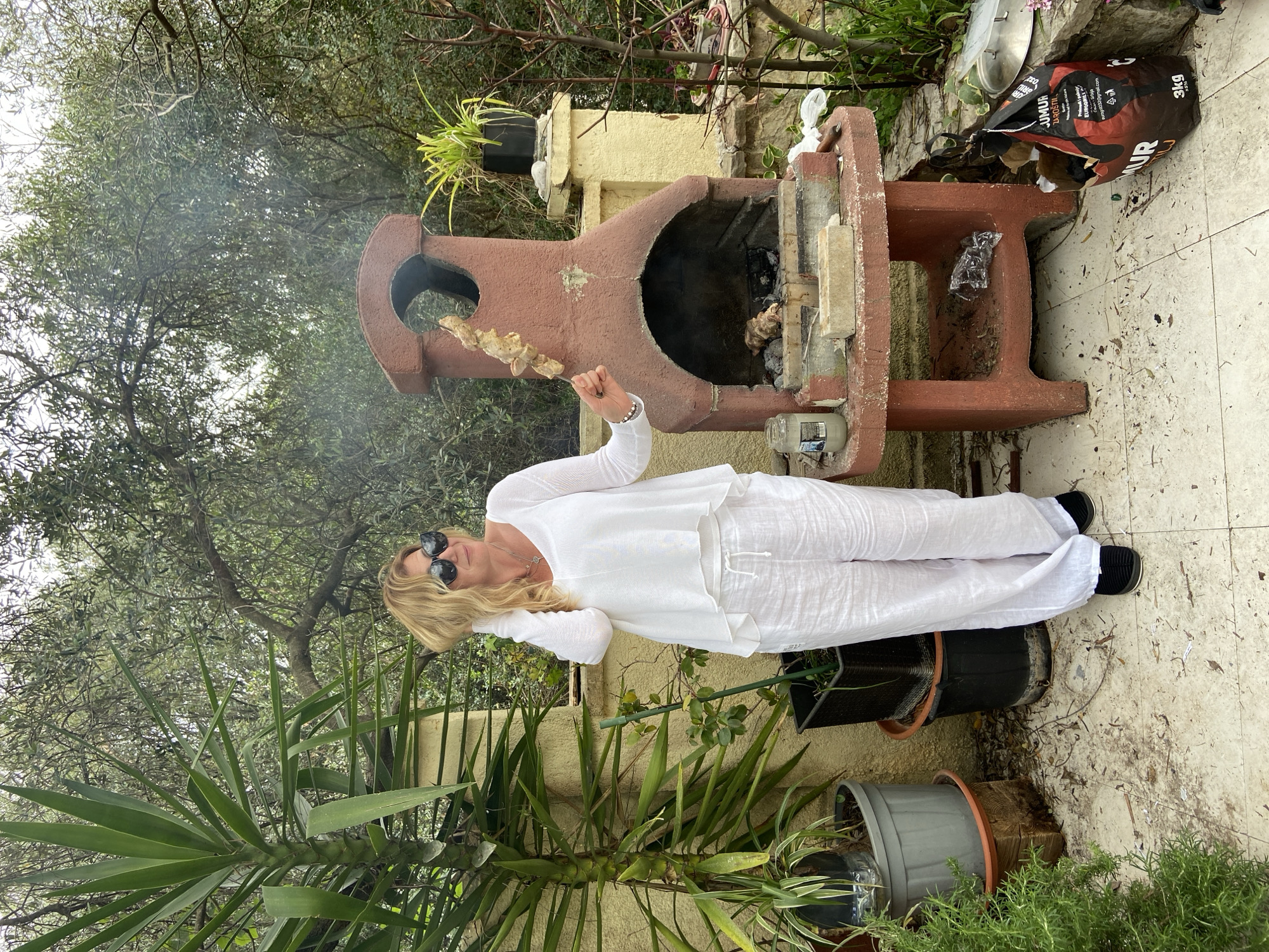 A person wearing a white outfit and sunglasses holds a skewer of food while standing in front of a barbecue grill. The grill, made of a terracotta-colored material, is positioned on a tiled surface outdoors. To the left of the person, there is a large leafy plant in a pot. Behind the grill, there is a bag of charcoal and additional greenery. The environment suggests a garden or patio setting, and the trees in the background imply a natural or residential area. The image was most likely taken in the city of Budva, Montenegro.