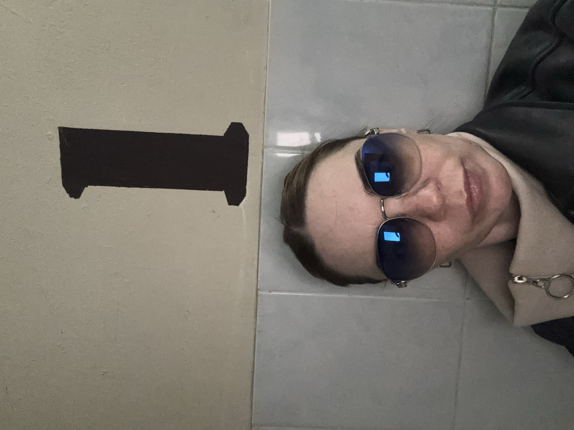 The image shows a person's face and shoulder, wearing sunglasses, a beige shirt, and a black jacket. The person is lying down next to a wall that appears to be tiled. On the wall is a dark, rectangular shape with protrusions on either side. The location is labeled Unknown, Russia.