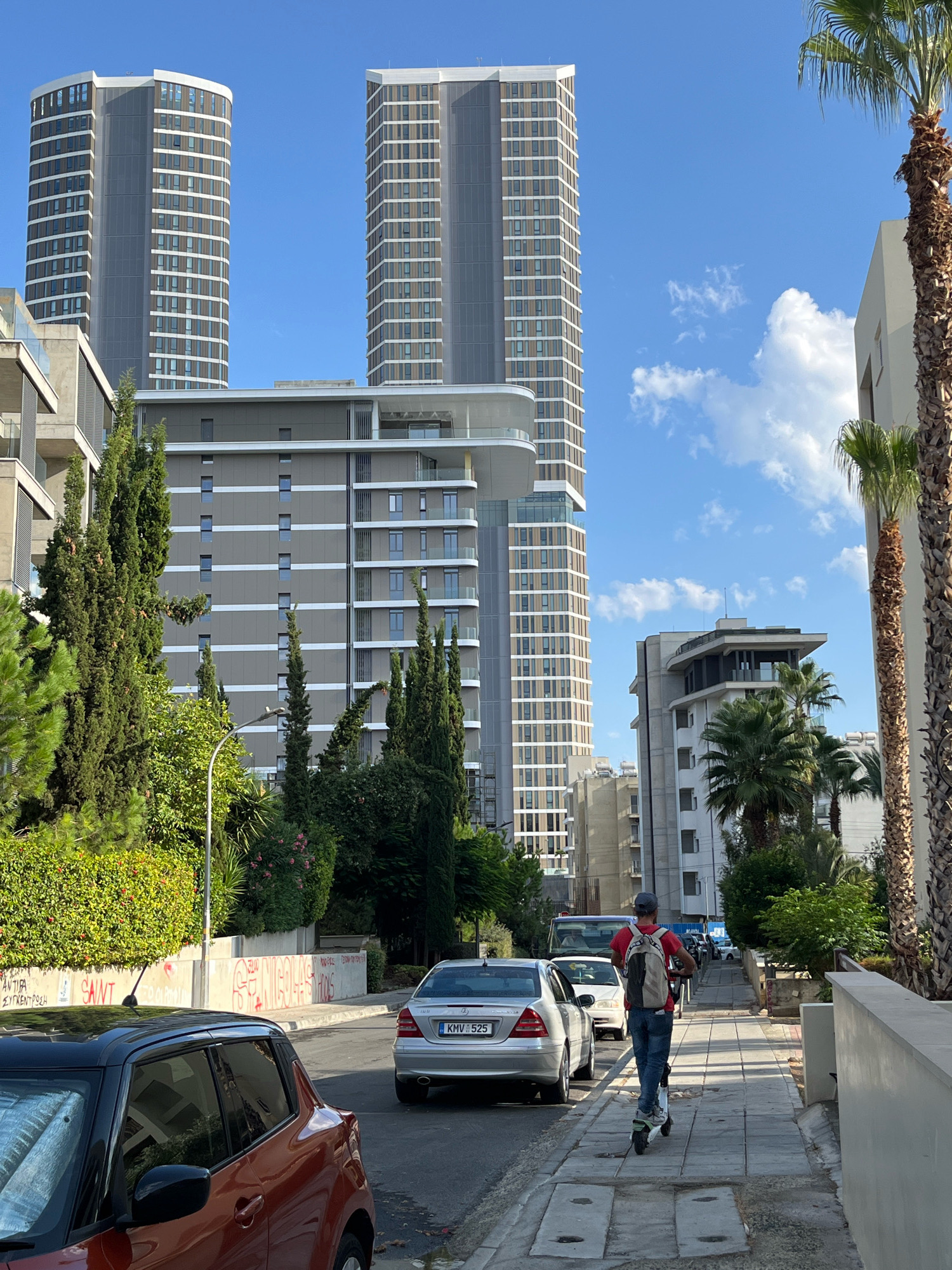The image depicts a street scene in Limassol, Cyprus, on a clear day with a blue sky and scattered white clouds. In the foreground, an orange car with a black roof is parked on the left side of the road. To its right, a silver Mercedes sedan is driving away from the camera, followed by a person riding a white electric scooter on the sidewalk. This individual is wearing a red t-shirt, blue jeans, and a backpack.
The street is lined with buildings of varying architectural styles. Two tall, modern skyscrapers with many windows dominate the background. In the middle ground, a grey building with balconies and white horizontal accents is visible, along with a lower, white building with a flat roof and prominent windows. Lush green trees and bushes line the street and sidewalk, with palm trees also present. Graffiti is visible on a low concrete wall on the left side of the road, with some text and red spray paint. The sidewalk is paved with grey square tiles. The overall impression is of a contemporary urban environment integrated with significant greenery.