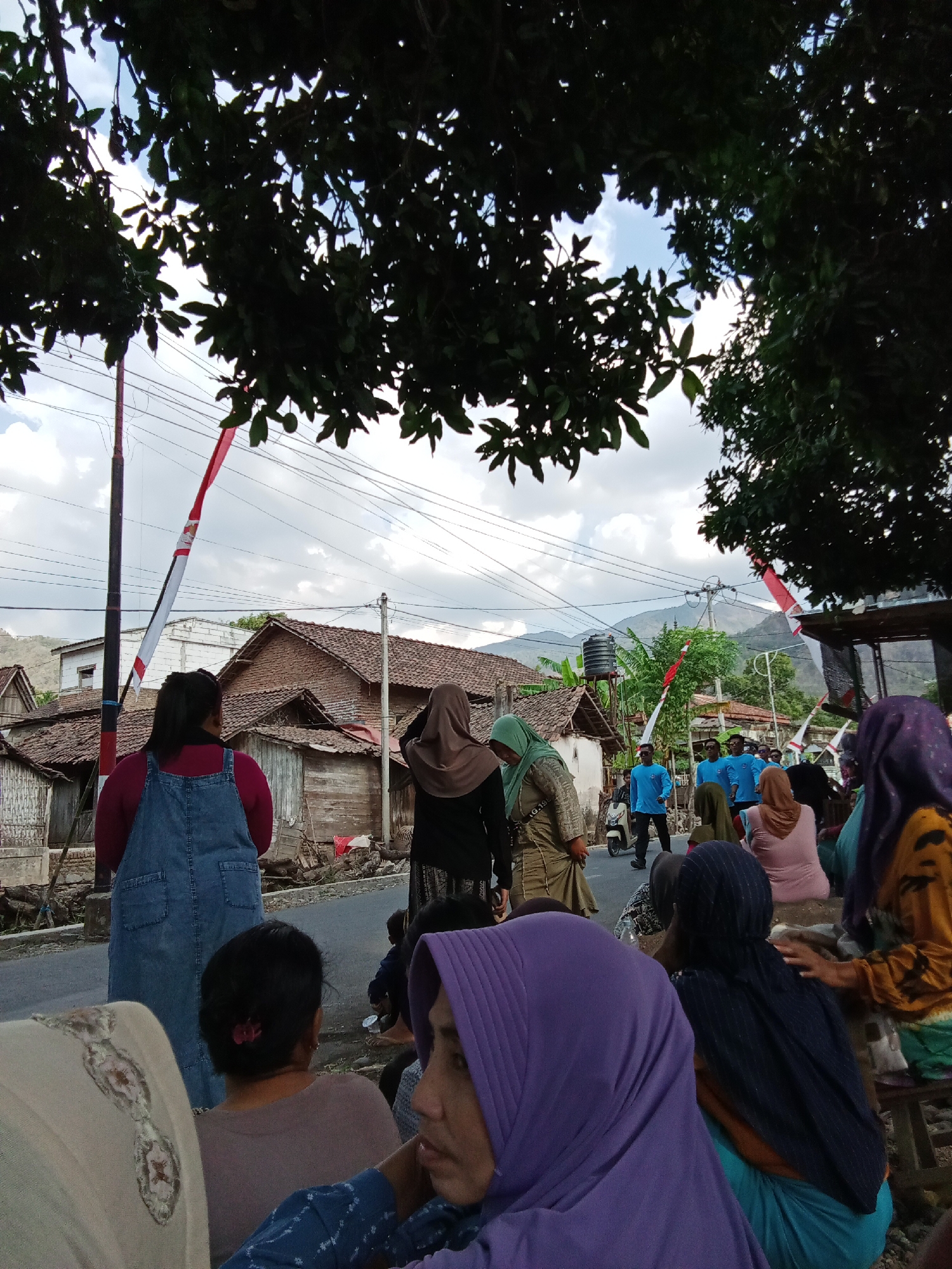The image depicts a street scene in Trebungan, Indonesia. Several people are present, many of whom are wearing headscarves. In the foreground, a woman in a denim overall is seen from behind, facing away from the camera. Adjacent to her, another woman in a brown hijab is also visible. The background shows a cluster of houses with tiled roofs and wooden elements, typical of Indonesian architecture. A red and white striped flag is tied to a pole, suggesting a local event or celebration. Several people in bright blue shirts are walking down the street, some of whom appear to be part of a group. The environment includes trees with dense foliage overhead, casting shadows on the scene, and a cloudy sky. Power lines and a water tank are also visible. The overall impression is a community gathering in a semi-rural Indonesian setting.