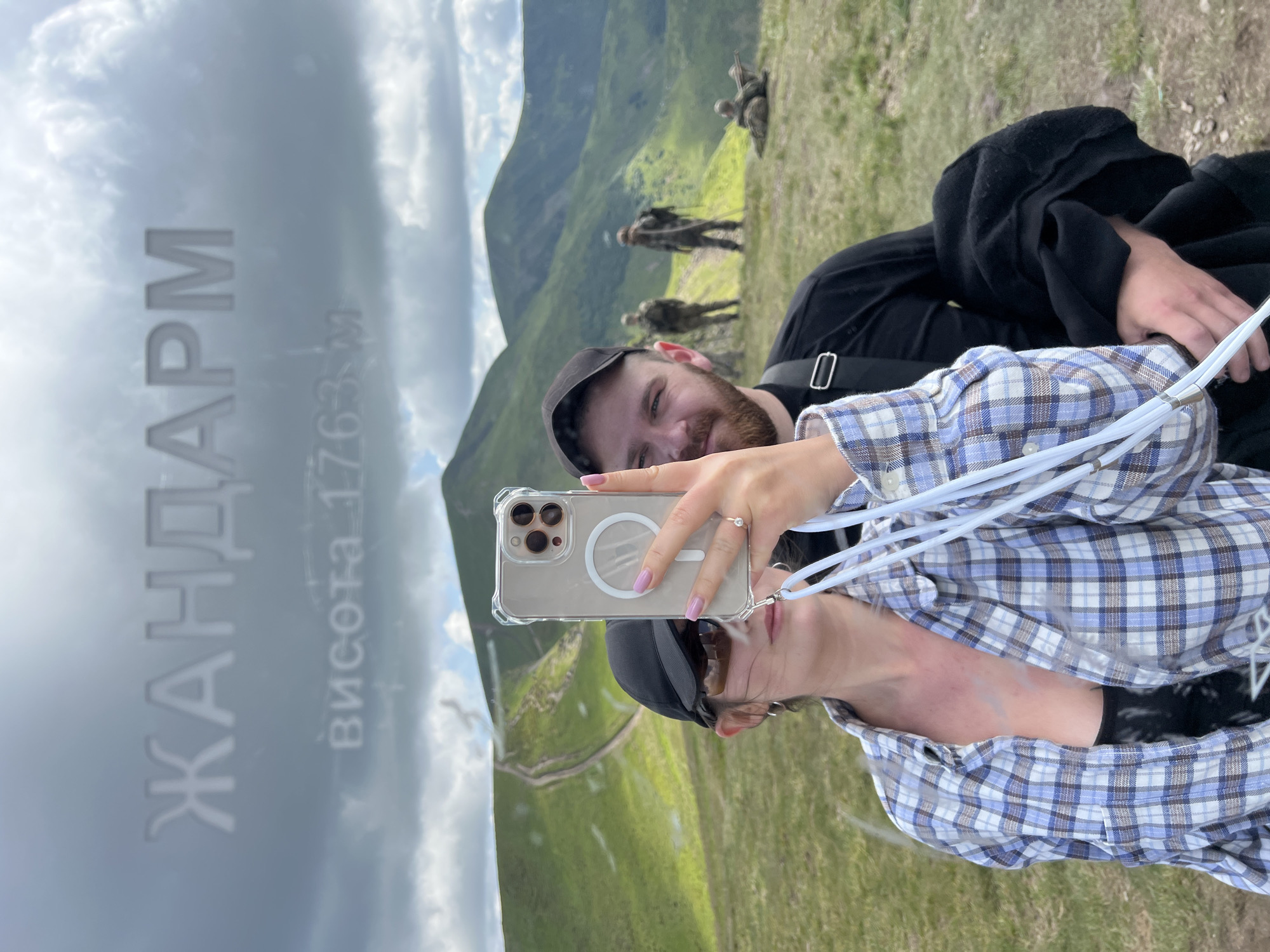 In Chorna Tysa, Ukraine, a couple takes a selfie on a mountain summit with a sign indicating "ЖАНДАРМ" and "висота 1763 м" (height 1763 m). The woman, in the foreground, wears a blue and white plaid shirt and a black baseball cap, holding up a smartphone in a clear case. Her companion, a man with a beard and a black t-shirt, is partially visible behind her. In the background, three individuals in military-style uniforms stand on the grassy mountainside, with a fourth person lying down. The landscape is characterized by rolling green hills under a cloudy sky.