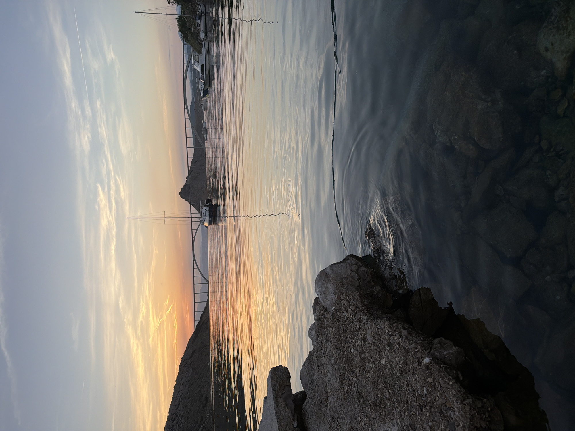 A wide-angle, low-angle shot captures a tranquil sunset over a body of water, likely a bay or strait, with a prominent arch bridge in the background. The sky displays soft hues of orange, yellow, and pale blue, with wispy clouds and a faint contrail from an aircraft. A rocky shoreline occupies the lower left quadrant of the image, with the water in the foreground appearing clear enough to reveal submerged rocks. Several sailboats and motorboats are moored or sailing in the middle ground, their masts and hulls reflected in the calm water. The arch bridge, a significant man-made structure, spans the water, connecting two hilly landmasses that frame the scene. The overall atmosphere is serene and picturesque, conveying a sense of peace and natural beauty at dusk.
