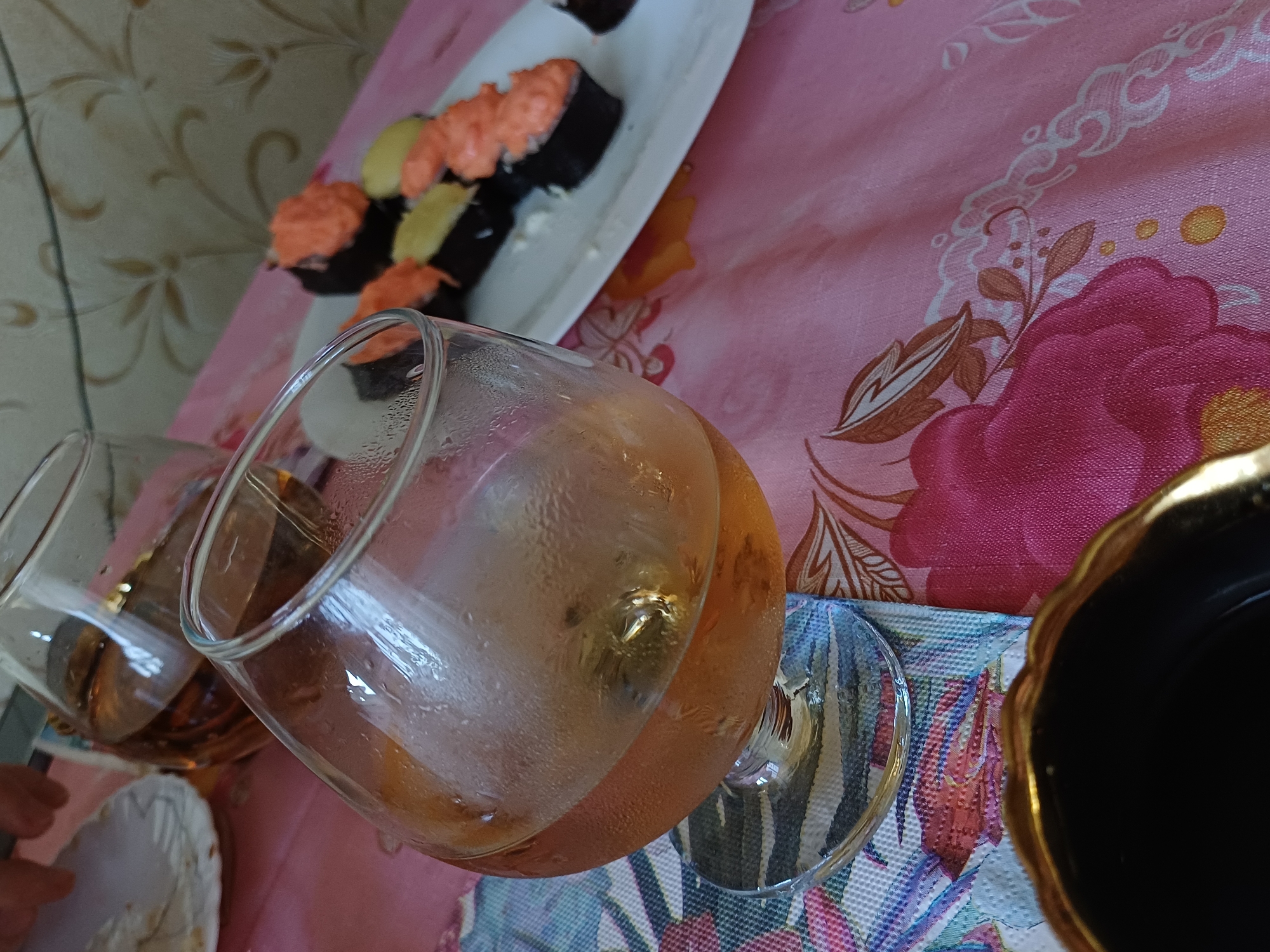 A still life image captures a close-up view of a table setting with a meal and drinks. In the foreground, a brandy snifter filled with a light brown liquid and ice sits on a patterned coaster, which rests on a pink tablecloth adorned with floral designs. To the right of the snifter, a white plate holds several pieces of sushi. In the background, another glass, partially obscured, contains a similar amber-colored beverage. The scene appears to be indoors, with a textured wall visible in the upper right corner. The lighting is soft, suggesting a casual dining or social gathering.