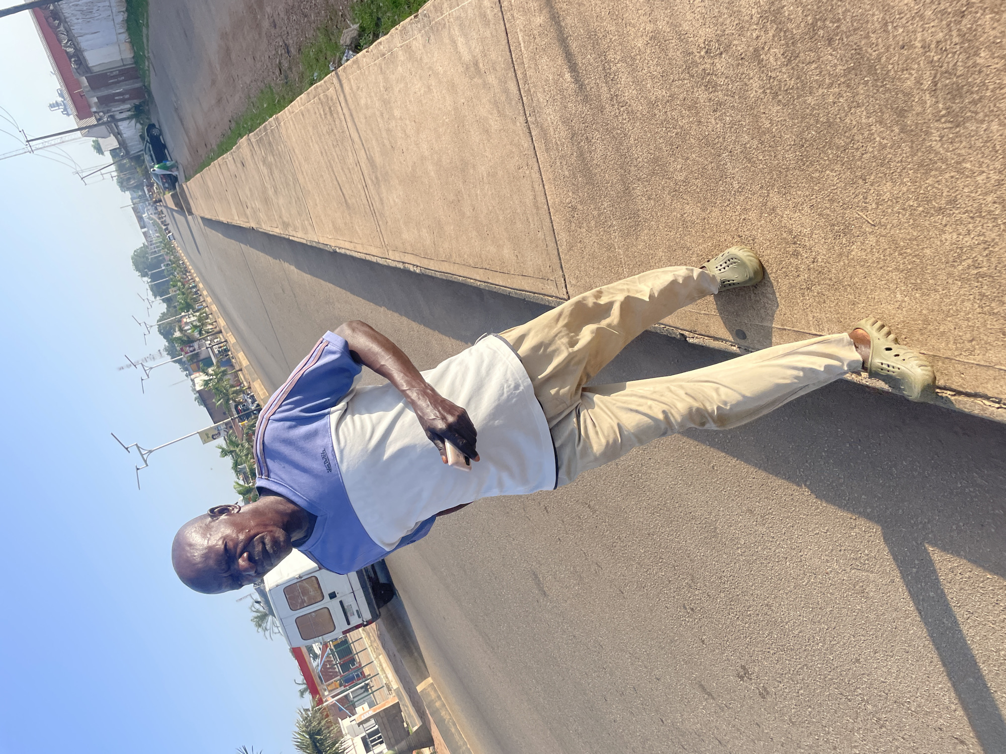 An outdoor, ground-level, medium shot captures a bald Black man walking along a concrete sidewalk next to an asphalt road. He is wearing a blue and white short-sleeved shirt, tan pants, and light green clogs. He is holding a dark-colored mobile phone in his right hand and looking towards the camera. The road has light traffic, including a white van and a black car. The environment appears to be a city street in Barakin Lerawi, Nigeria, with palm trees, streetlights, and buildings visible in the background. The sky is clear and blue, indicating daylight. The sunlight casts a long shadow of the man onto the sidewalk and road.