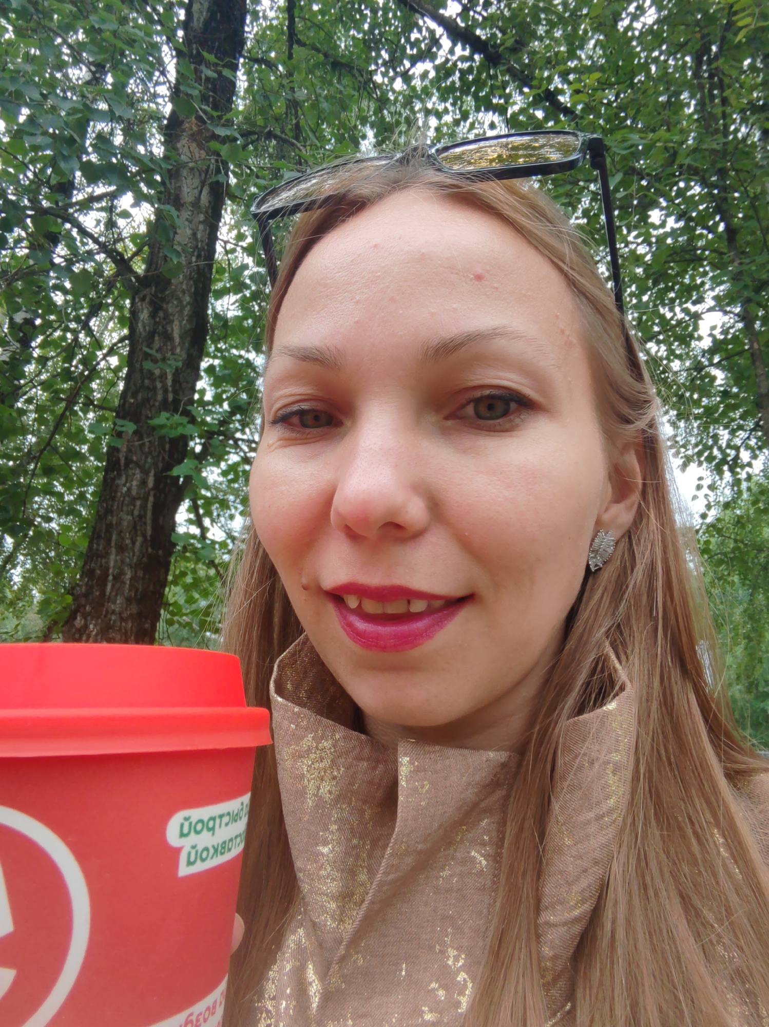 A woman with blonde hair, glasses on her head, and pink lipstick is holding a red cup with a white logo. She is wearing a gold-flecked jacket. She is outdoors, with trees and green foliage in the background. The image appears to have been taken in the city of Syktyvkar, Russia, given the Cyrillic text on the cup which reads "СКОРОСТЬ" (speed) and "НА ВКУС" (to taste).