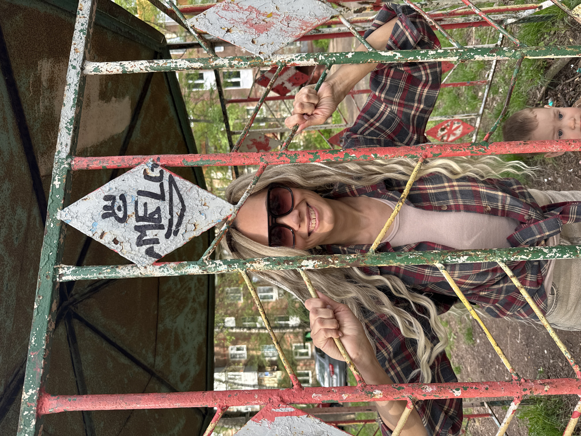 The image shows a woman with blond hair and sunglasses smiling at the camera. She is holding onto the metal bars of a structure painted in red, green, and white. The metal structure contains diamond-shaped elements, one of which has the word "MELC" written on it. Behind the structure, there is a blurry view of trees and buildings, potentially residential. In the background, the face of a child can be partially seen. Based on the presence of older, possibly playground-type metal structure and apartment blocks, this may be a location in a residential area. The name "MELC" written on the structure would suggest that this image was taken at Rusanovo in Russia. The photo seems to be taken looking up at an angle, as if the person taking the picture is below the woman and the metal structure.
