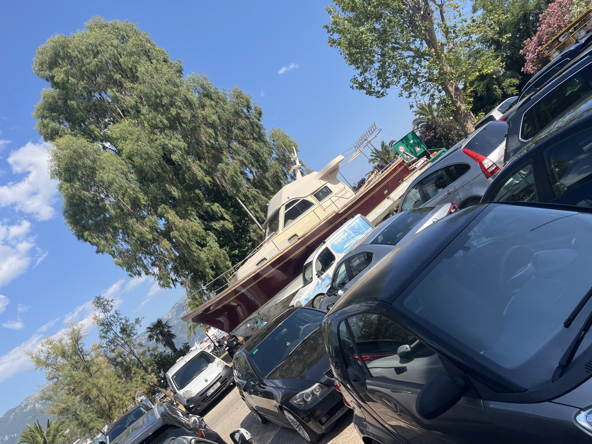 In Budva, Montenegro, a large, maroon and cream-colored boat rests on a trailer, seemingly in a parking lot or staging area. The boat, with the marking "TY 2" visible on its side, is positioned at an angle, partially obscured by several parked cars. The cars are of various models and colors, including black, silver, and white. Some of the cars have their license plates visible, with one reading "GU DK 681".
The scene is bathed in bright sunlight, suggesting it is daytime, likely midday or early afternoon, given the clear blue sky with a few scattered white clouds. Lush green trees, including a large, prominent tree with feathery leaves on the left, and other foliage, surround the boat and cars, indicating a setting with natural elements. In the background, distant mountains can be seen. The overall impression is a sunny, outdoor scene in a coastal or near-coastal environment. There are no people visible in the immediate foreground or on the boat, but the presence of multiple parked cars suggests a public area. A green sign with white text and a circular symbol is also visible on the trailer supporting the boat. The overall atmosphere is one of stillness, with the boat temporarily out of the water and surrounded by vehicles.