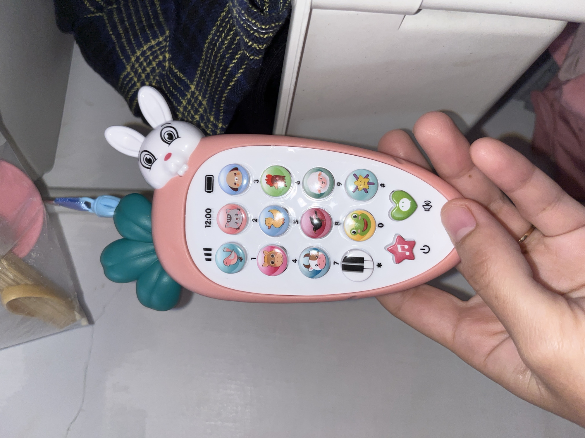 A right hand, adorned with a thin gold ring on the ring finger, holds a pink, carrot-shaped infant's learning toy in an indoor setting in Lhokseumawe, Indonesia. The toy features a white rabbit head with black-outlined eyes and a pink nose protruding from its top, positioned above three teal-colored, leaf-like structures. The toy's central screen area is white, displaying "12:00", three full signal bars, and a full battery icon.
Below the screen, a grid of twelve circular buttons is arranged, primarily depicting cartoon animals. From left to right, top to bottom, these include a light blue bird (above the number '1'), a pink cat (above '2'), a light blue dog (above '3'), a pink cow (above '4'), a light blue duck (above '5'), a green rooster (above '6'), a button with piano keys (white and black, above '7'), a pink button with a black bird (above '8'), and a light pink sheep (above '9'). The bottom row features a red star with a white musical note (positioned above a power symbol), a yellow-green frog face (above the number '0'), and a light blue starburst design (above a hash symbol '#'). A green heart button with a white speech bubble is visible to the right, above a volume icon.
The hand holds the toy by its lower left side, with the thumb visible on the left edge and other fingers wrapped around the bottom. The background consists of a light grey wall with a hairline crack in the upper left, a dark blue fabric with a yellow plaid pattern on the right, and a white shelf or surface extending horizontally behind the toy. In the upper left corner, a transparent plastic bag containing light brown, woven material and a pink plastic object, along with a blue plastic clip, is partially visible.