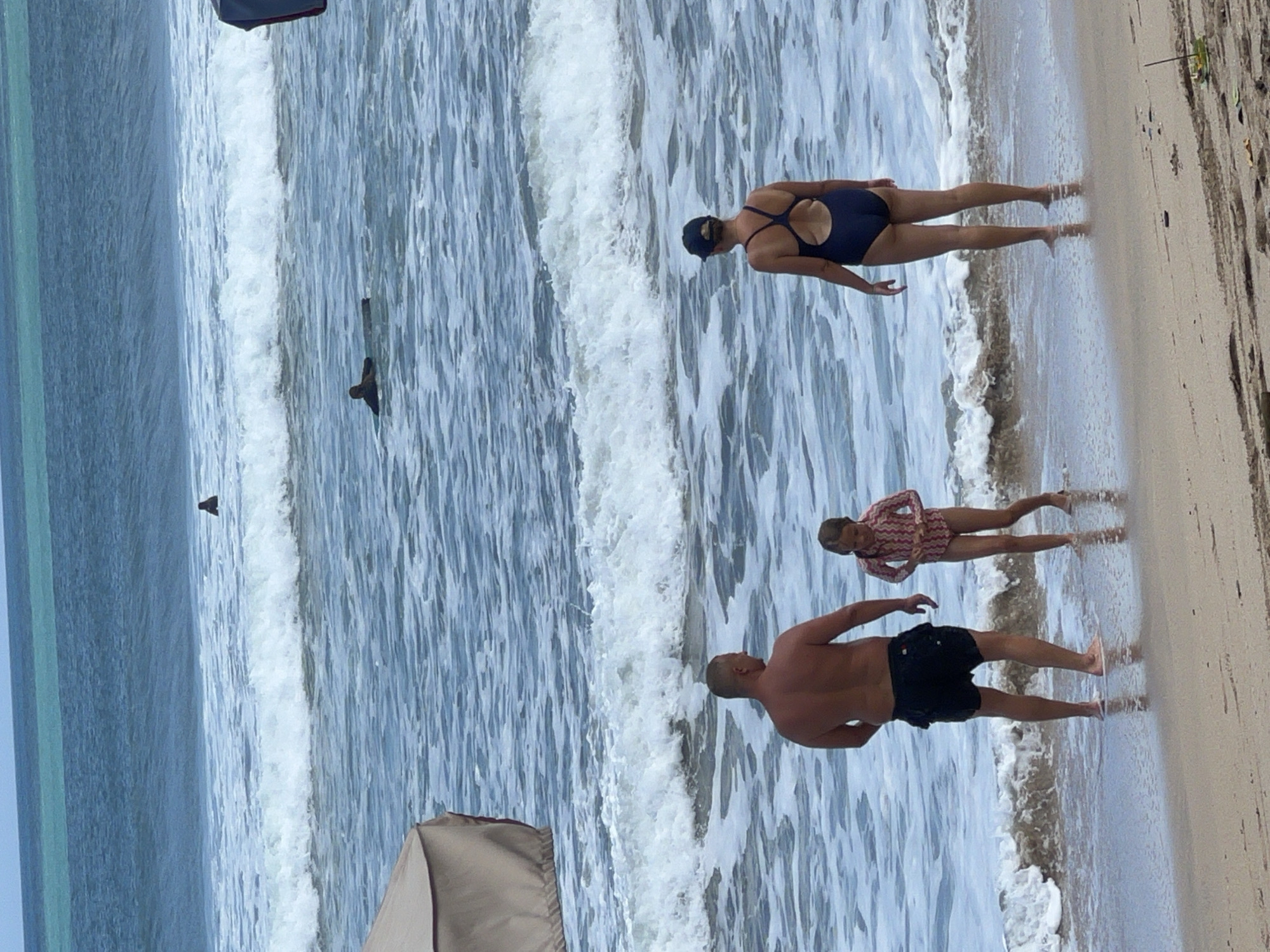This is an eye-level, medium shot of a family at the beach in Kuta, Indonesia. In the foreground, a man with no shirt and black shorts stands with his back to the camera, facing the ocean. To his left, a young girl in a long-sleeved patterned swimsuit stands facing the man, her hands at her sides. To the man's right, a woman in a dark blue one-piece swimsuit and a dark baseball cap stands with her back to the camera, facing the ocean. The ocean has moderate waves and several surfers can be seen in the distance. A part of a tan beach umbrella is visible in the upper left corner of the image. The sand is wet where the waves are reaching the shore. The sky is a clear blue with a hint of turquoise water visible in the distance.