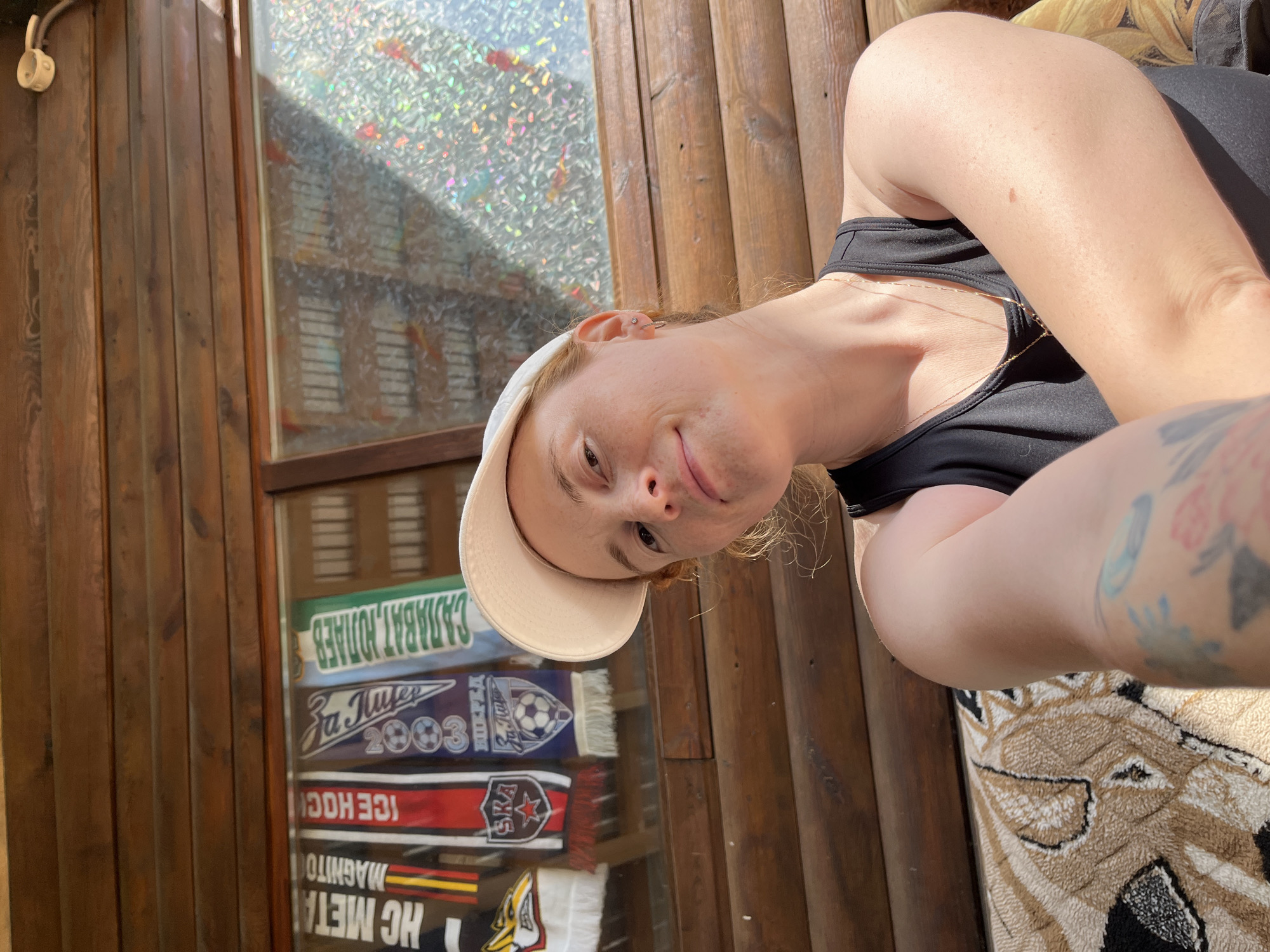 A woman with fair skin and reddish-blonde hair, wearing a black tank top and a light-colored baseball cap, poses for a selfie in front of a wooden structure with a glass-fronted display case in the city of Chaikovsky, Russia. The display case contains various sports scarves, including one with "CARABAJAT, HOTAEB" and another that says "ICE HOCK." The woman has a tattoo on her upper left arm. The background shows wooden paneling and a reflective glass surface behind the display case. She is sitting on a patterned surface.