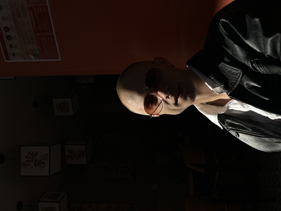 A bald male individual, appearing to be in his 20s or 30s, is depicted in a medium-close shot, seated within what appears to be an Asian-themed restaurant or cafe in Paris, France. He faces the camera with a faint smile, wearing amber-tinted aviator sunglasses, a white collared shirt, and a black leather jacket. A small silver earring is visible in his left ear. Strong, direct sunlight illuminates the left side of his face and upper torso, creating a sharp contrast with the deeply shadowed right side.
The background on the left is predominantly dark, revealing several square paper lanterns suspended from the ceiling. These lanterns are adorned with red Asian characters, with "平安喜樂" (peace and joy) clearly visible on one. A vague, shadowy form of another person or object is discernible in the far left background. The floor beneath the dark area appears tiled.
The right side of the frame features a bright orange wall. A white-framed rectangular poster, functioning as a menu, is affixed to this wall. The menu text is in French, listing dish names such as "NOUVELLE RANDONNÉE" and "SOUPE DE MOULE," along with prices. The combination of the French menu and Asian decor confirms the location is an establishment in Paris, France. The scene is illuminated by natural light, casting pronounced shadows.