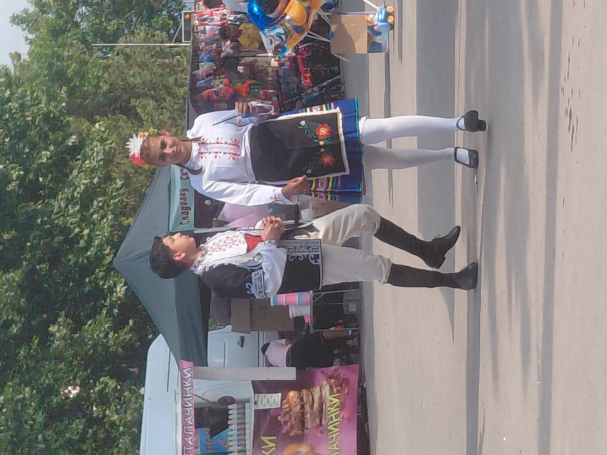 This image depicts an outdoor daytime scene in Unknown, Unknown, likely a market or festival. In the foreground, two young individuals dressed in traditional folk attire are prominently featured.
On the left, a male is seen in a white long-sleeved shirt with red and black embroidery on the collar and cuffs, a black sleeveless vest with white and light blue intricate patterns, white trousers, and tall black boots. He stands facing right, with his right arm bent near his stomach and his left arm bent lower.
On the right, a female is depicted wearing a white headpiece adorned with red, white, and other colored floral elements. She has a white long-sleeved blouse with red and white embroidery on the chest, a blue pleated skirt, a black apron with a colorful floral embroidered design (featuring red, orange, and green flowers), white opaque tights, and black low-heeled strap shoes. She holds a slender, dark object in her right hand and looks towards the viewer with a slight smile.
In the background on the left, a white food truck or stall displays a pink banner with the Cyrillic word "ПАЛАЧИНКИ" (Pancakes) and images of pancakes. Bottles are visible on a shelf of the stall. A dark green canopy tent and a white van are situated further back. To the right of the van, a person in a light pink shirt is bent over, possibly attending to items on a table. On the far right, a merchandise stall displays numerous colorful items, and a blue and yellow child's ride-on toy is visible on the ground along with a cardboard box. The ground is a light-colored paved surface. The upper background is dominated by lush green trees, indicating an open-air environment under a bright sky.