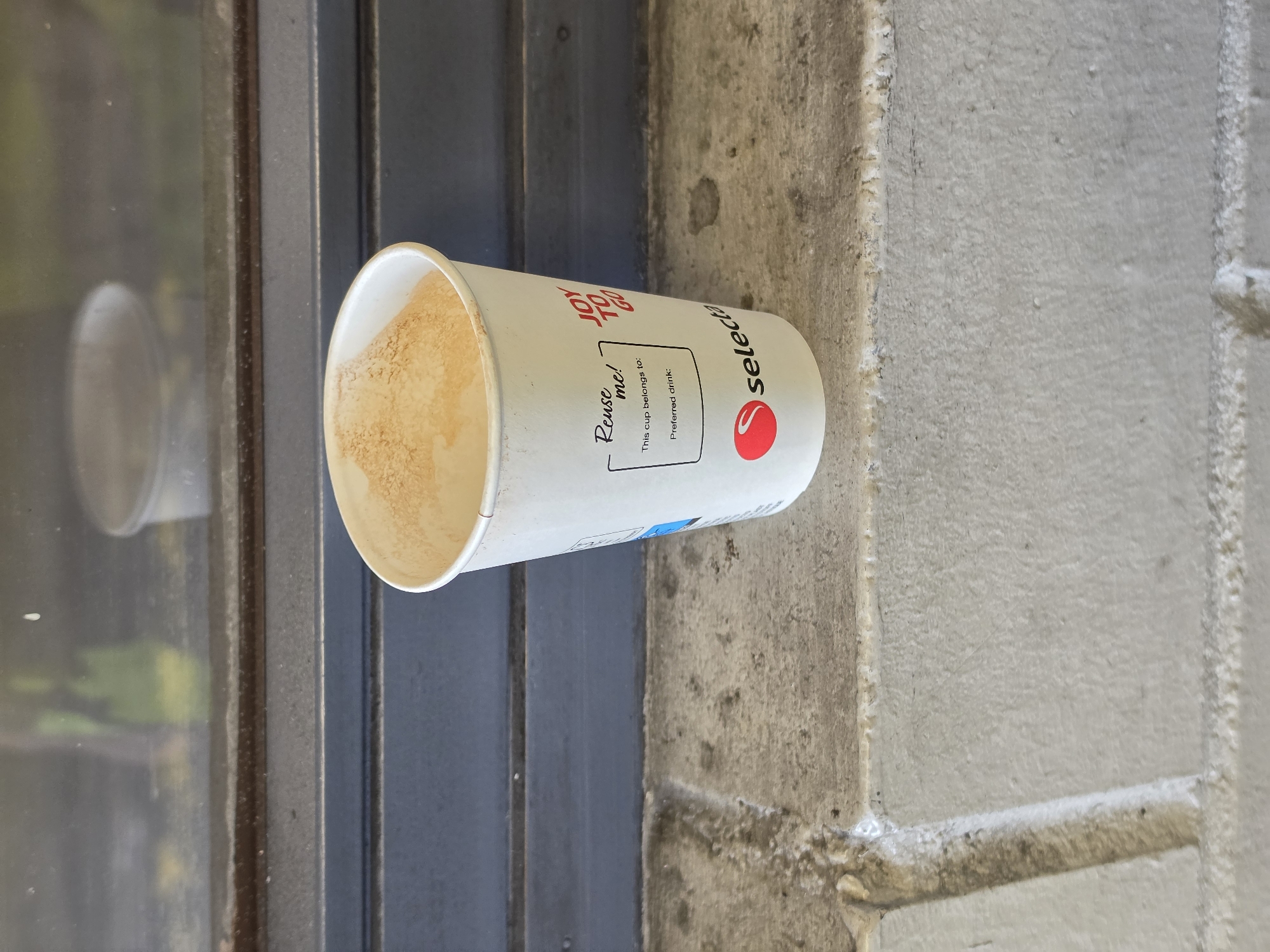 A disposable white paper cup, partially filled with a light brown, foamy substance, rests on a textured, light gray concrete ledge. The cup has a logo and text printed on it, including "Reuse me!", "This cup belongs to:", "Preferred drink:", and "Selecta" with a red circle logo. The background consists of a dark gray, slatted surface and a window with reflections, suggesting an urban outdoor or semi-outdoor setting, possibly near a building. The scene is well-lit, with natural light casting soft shadows. No people are visible in the image, and no specific location within Frankfurt, Germany can be determined.