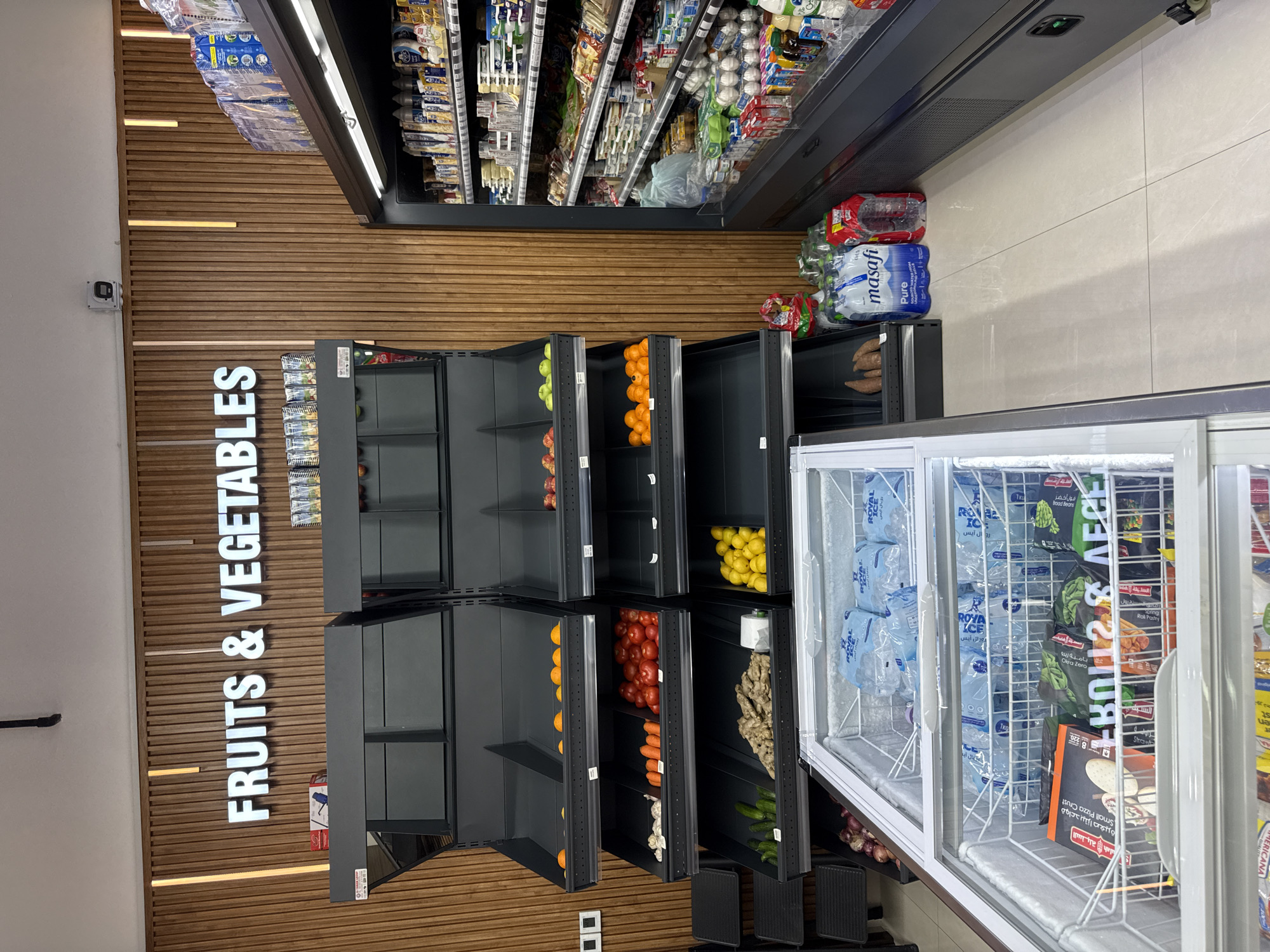 The image shows the interior of a grocery store, possibly in Dubai, United Arab Emirates. The foreground features a large, open chest freezer filled with bags of ice and frozen food items. Beyond the freezer, rows of shelving display various produce, including oranges, tomatoes, carrots, cucumbers, ginger, and lemons. Some shelves are empty, while others hold a limited selection of fruits and vegetables. To the right, a refrigerated display case is stocked with dairy products, drinks, and other packaged goods. The store's walls are decorated with vertical wooden paneling, and a prominent white sign reads "FRUITS & VEGETABLES." The overall impression is of a well-lit retail environment with a focus on fresh and frozen produce.