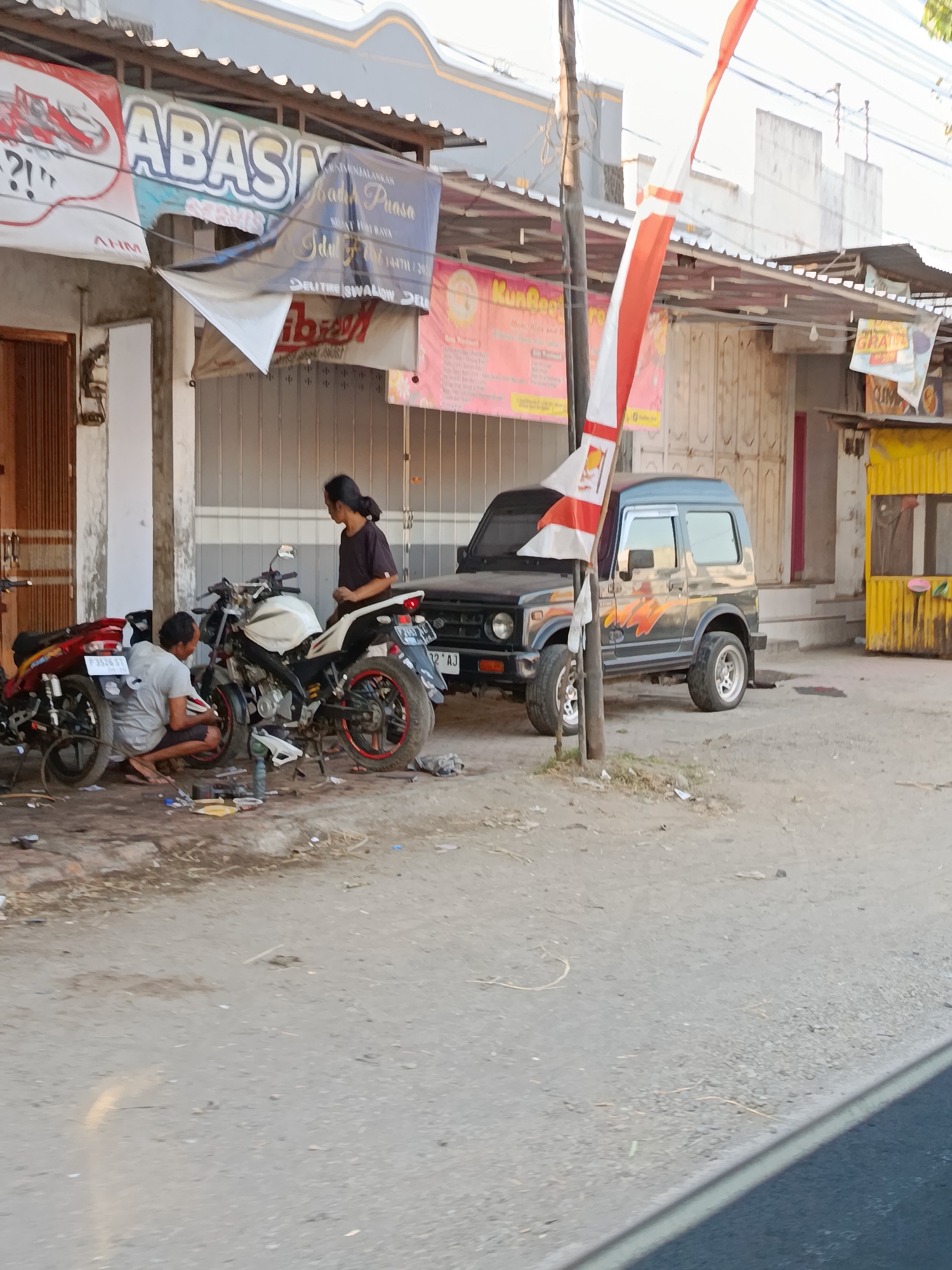 The image shows a street scene in Wonosari, Indonesia, likely in front of a commercial establishment. Two men are working on a white motorcycle parked on a dirt and gravel ground. One man is kneeling and appears to be working on the front wheel of the motorcycle, while the other man stands beside it, looking down. To the left, a red motorcycle is parked. In the background, there is a dark-colored SUV parked. A building with various signs is visible behind the motorcycles and SUV. One sign, partially obscured, has large blue lettering. Another sign in pink features yellow text. A tall pole with Indonesian flags, red and white, is positioned near the SUV. The ground is uneven and dusty, with some debris scattered around. The lighting suggests it is daytime, with sunlight casting shadows. The overall scene depicts a roadside repair area or a place of business servicing vehicles.