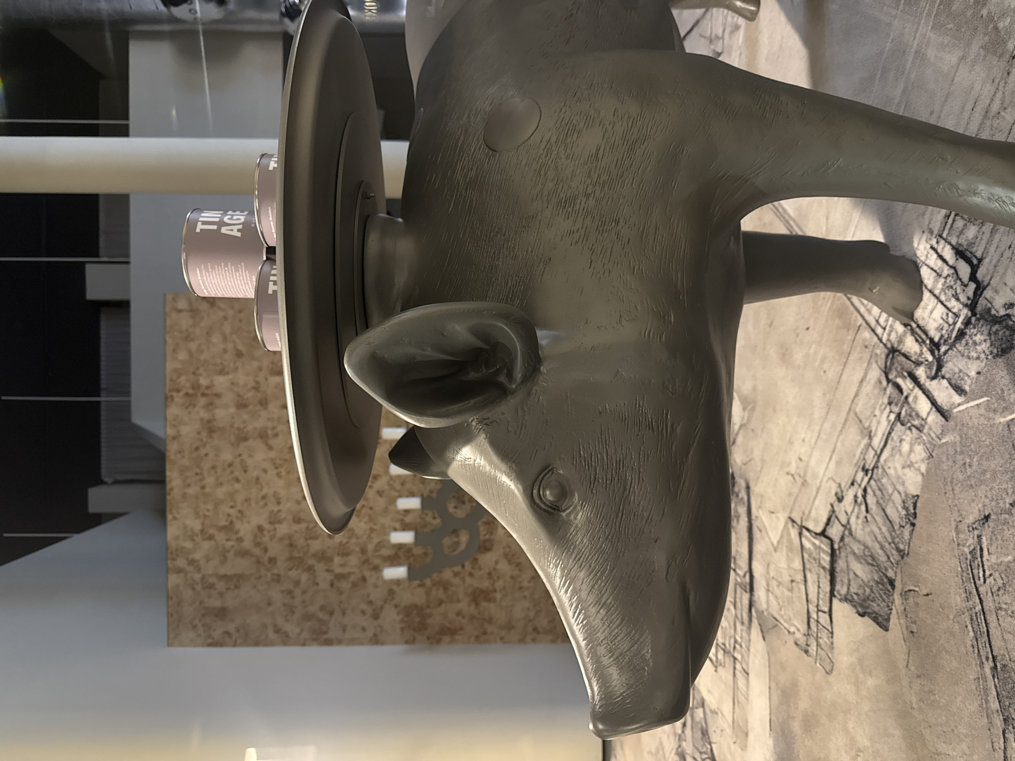 A large, stylized statue of a quadruped animal, resembling a pig or a similar domesticated ungulate, is the central subject, rendered in a matte, dark grey finish. The statue is positioned horizontally as if lying down, with its head facing the right side of the frame. Its surface exhibits subtle linear textures that suggest fur or bristles, and its eyes are depicted as simple raised circles.
Mounted onto the animal's back is a flat, dark circular disc, from which several small, light pinkish-brown cylindrical containers (likely cans or jars) protrude upwards. The words "TIN AGE" are clearly visible, split across two lines, on the side of these containers.
The scene is set indoors, characteristic of a museum, art gallery, or exhibition space. The background features a large, light-colored graphic print on a wall or floor, which appears to be an intricate architectural drawing, blueprint, or map. Behind the animal's head, there is a wall with a mottled, light brown pattern, and further back, white cylindrical columns and darker architectural elements are visible. Abstract white shapes can also be faintly discerned on the brown patterned wall.
There is no human activity depicted. The media captures an art installation, notable for its unusual composition of an animal figure serving as a base for industrial-style containers. The monochromatic color palette of the statue contrasts with the subtle hues of the background and containers, emphasizing the sculpture's form and the symbolic interaction between its elements. The arrangement suggests a conceptual art piece.