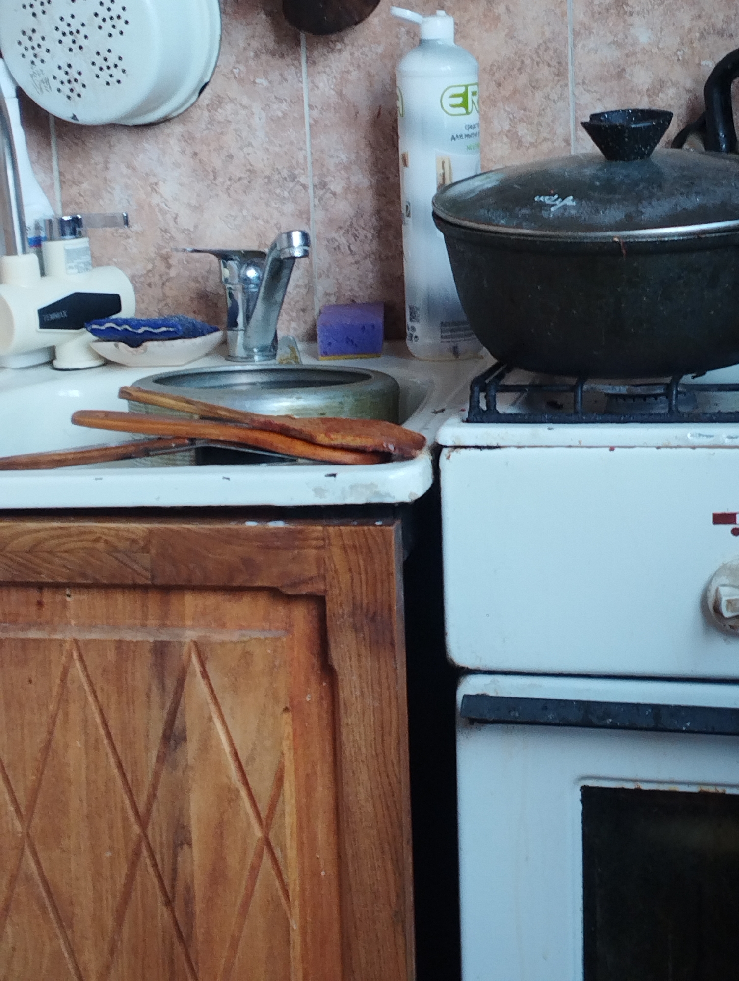 The image depicts a cluttered kitchen scene, likely within a residential setting in Bishkek City, Kyrgyzstan. In the foreground, a wooden cabinet with a carved diamond pattern is visible. To the right of the cabinet is a white stove. On top of the stove rests a large, dark pot with a lid and a black knob. Next to the stove, a white sink is present, with a faucet and a soap dispenser. A blue sponge and a soap dish with a blue sponge are also on the edge of the sink. Two wooden spoons are lying across the sink. In the background, a white colander hangs on the wall, and a bottle of "ER" brand liquid soap stands near the sink. The wall behind the objects appears to be tiled with a textured, beige material. The overall impression is one of a functional, lived-in kitchen.