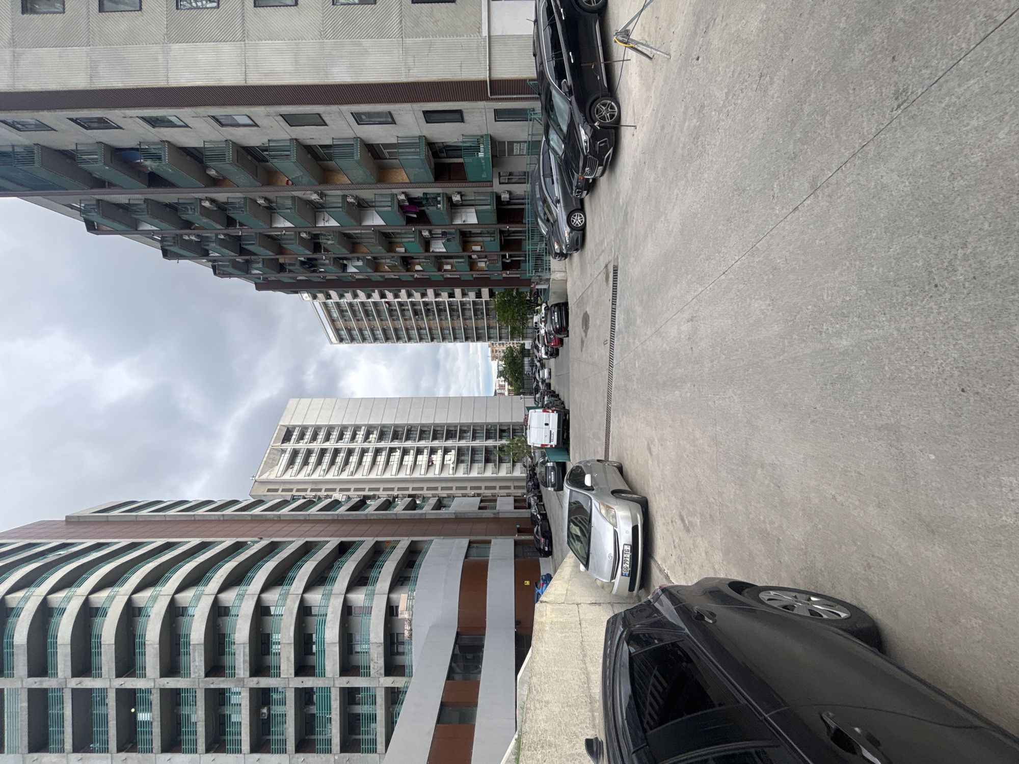 The image shows a street scene in Tbilisi, Georgia, flanked by tall apartment buildings on both sides. Cars are parked along the right side of the road. The buildings vary in design, one featuring rows of balconies and another with curved architectural elements. The sky is overcast. In the distance, more high-rise buildings are visible.