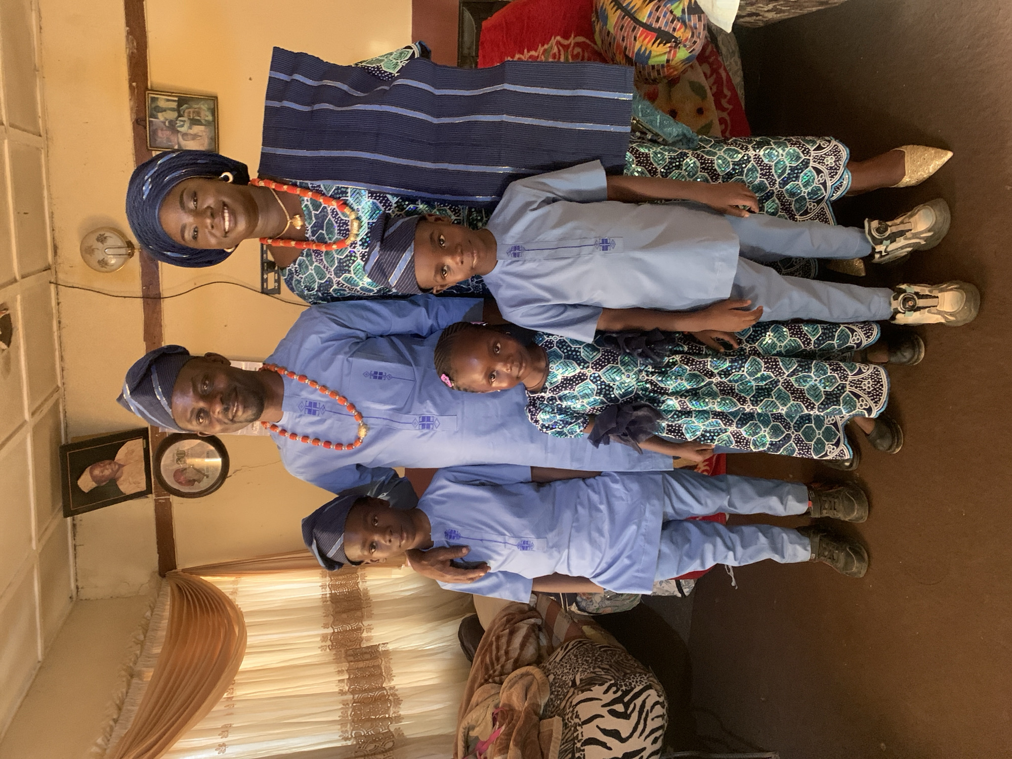A family of five poses for a photograph indoors in Rafin Bauna, Nigeria. The family includes a mother, father, two sons, and a daughter. The mother stands at the top left of the frame wearing traditional Nigerian clothing of blue, black, and green with a matching head wrap and red coral jewelry. The father stands below the mother, wearing a light blue traditional shirt with similar red coral jewelry and a blue head covering. Their sons wear matching light blue outfits with blue head coverings. The daughter is positioned between the father and elder son, wearing a green and blue patterned top similar to the mother's clothing. The family is arranged vertically, facing towards the viewer. The background includes a room with cream-colored walls, curtains, and framed photographs on the wall. The floor is brown. A patterned fabric is visible behind the mother and sons.