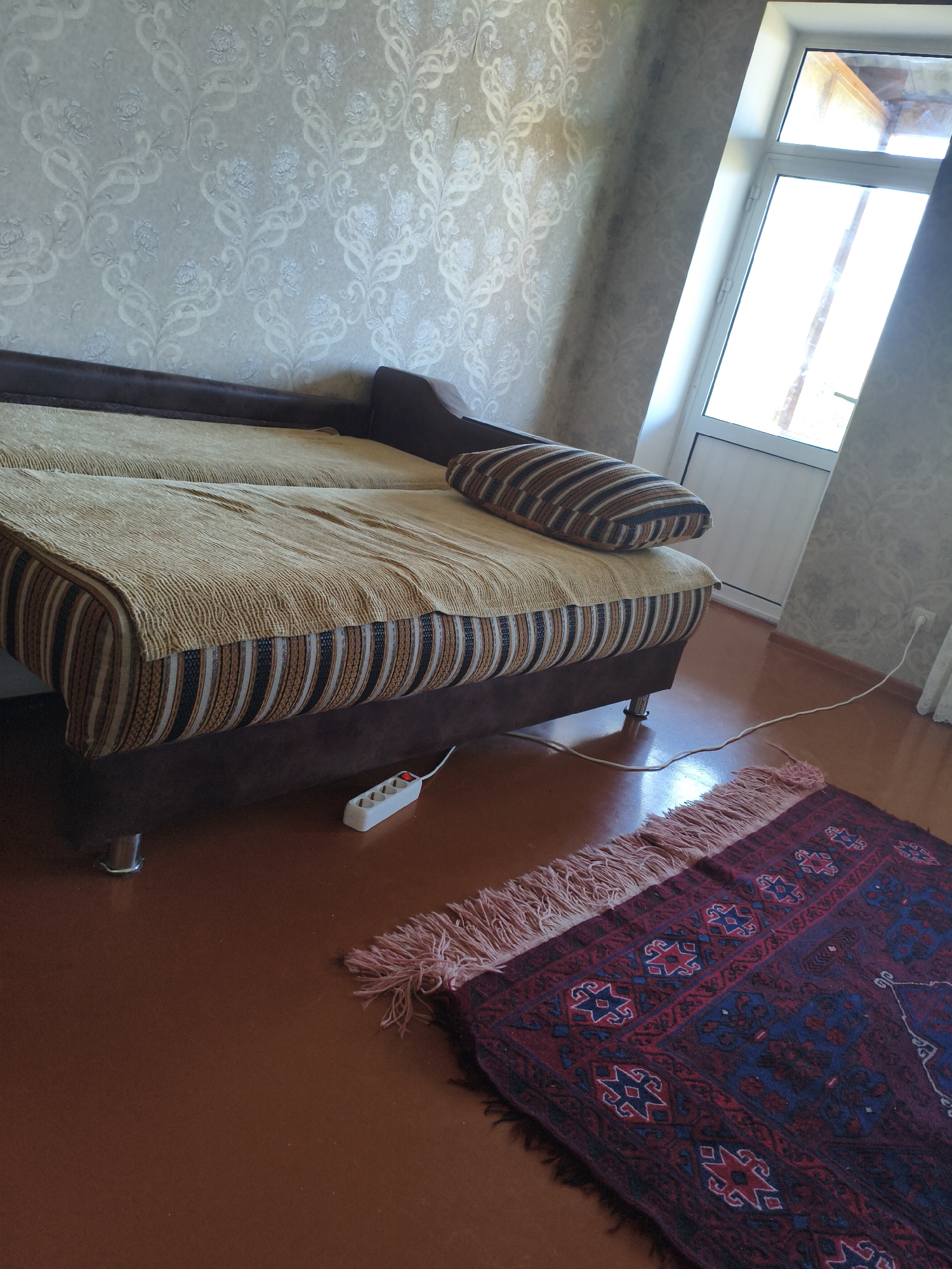 The image depicts an interior scene, likely a bedroom or living area, featuring a couch, a rug, a window, and a wall with patterned wallpaper. The couch is covered with a beige throw blanket and has a striped pillow. A white power strip with a red switch is plugged into an outlet near the couch, with a white cord running along the floor. A small, intricately patterned rug with a fringed edge is placed on the brown wooden floor. A large window with a white frame allows natural light to enter the room. The wallpaper has a light, floral pattern. The overall impression is of a lived-in space, possibly a domestic dwelling. The location is identified as Derbent, Russia.