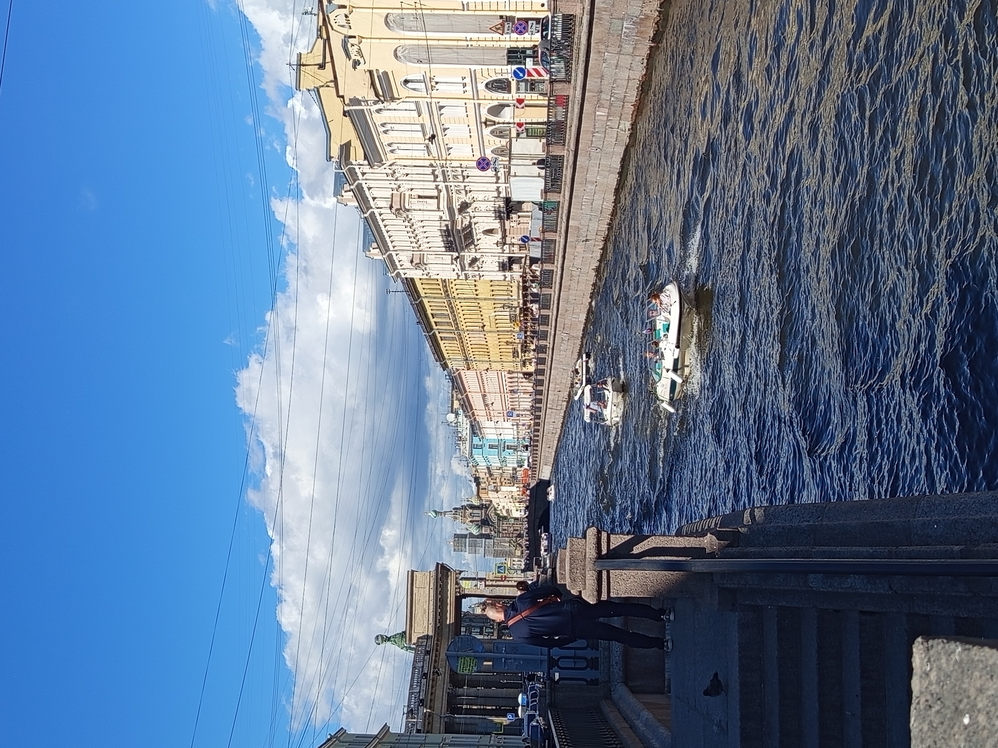 The image captures a sunny day scene along a canal in Saint Petersburg, Russia. The canal is flanked by ornate buildings with pastel facades, characteristic of the city's architecture. Several boats are navigating the dark blue water, some appearing to be tour boats with passengers. On the left side of the canal, a person stands on a stone embankment, looking out at the water. The sky is a vibrant blue with scattered white clouds, and power lines are visible crisscrossing the upper portion of the frame. In the distance, a prominent domed building, likely a church, rises above the cityscape. The overall impression is of a lively, picturesque urban environment.