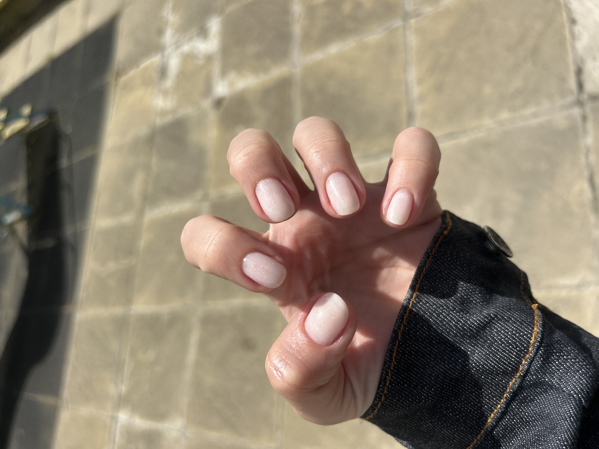 A close-up photograph shows a person's left hand against a blurred backdrop of beige pavement. The hand is slightly curled with the fingertips pointing upward. The person's nails are short and rounded, painted with a pale, possibly pearlescent, nail polish. The subject is wearing a dark blue denim jacket, the cuff and a button visible on the right side of the frame. The image is taken in natural daylight. The location is outside, evidenced by the paved surface. I cannot identify the city as Sofia, Bulgaria, without additional information.