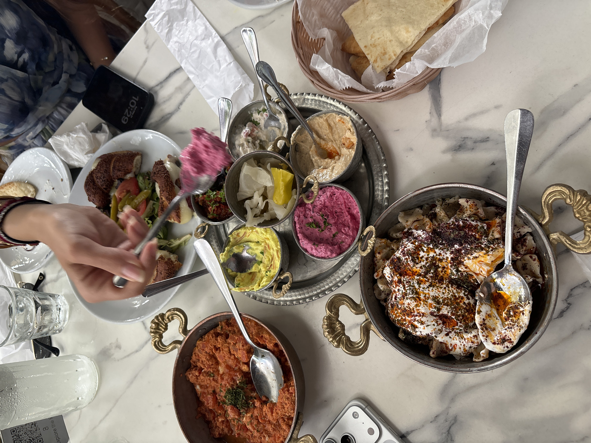 A top-down view captures a dining table scene with a variety of foods presented on a white marble-patterned surface. A woman's hand is visible, manipulating a spoonful of a magenta-colored substance towards a plate containing slices of sesame-crusted bread interspersed with salad vegetables. The plate sits near a black smartphone displaying a time of 10:29.
A silver tray holds several small, ornate metal dishes filled with various dips, including a beige hummus-like spread, a white, creamy mixture, a red paste, a yellow spread, and two different shades of magenta. Each dish has a spoon inserted for serving.
Adjacent to the tray are two more metal dishes with decorative handles. One contains a tomato-based mixture, while the other appears to hold a dumpling-like dish topped with a white sauce and sprinkled with dark spices. A spoon rests in this dish as well. A basket filled with flatbread sits at the top of the frame.
A clear water bottle and a glass of water are positioned to the left. The table also has a white smartphone with a camera lens visible. The scene appears to be a restaurant setting, possibly in a city known for diverse culinary experiences, like Miami, United States of America.