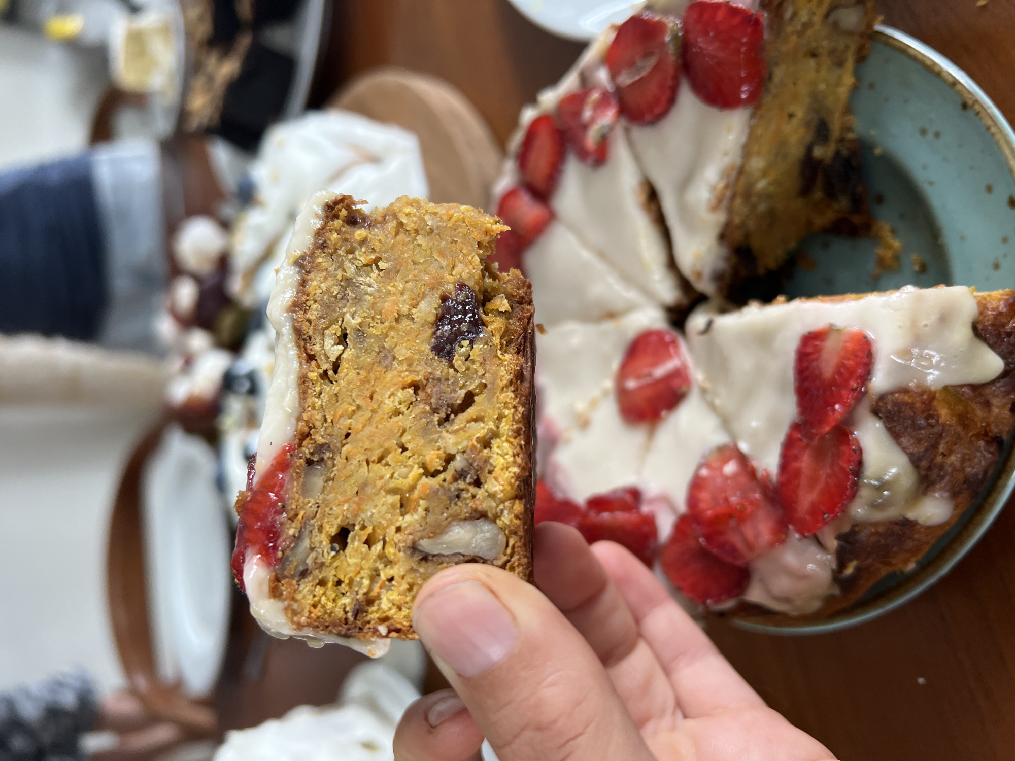 The image shows a slice of carrot cake being held in a person's hand. The cake slice has a creamy white icing with a reddish jam-like substance dripping down one side. The cake itself contains visible shredded carrots, nuts, and possibly raisins. In the background, there is a cake with a similar creamy icing, topped with sliced strawberries, on a plate. Other plates and food items are visible in the periphery, suggesting a meal or gathering. The location, Singakerta, Indonesia, is not discernible from the image content alone.
