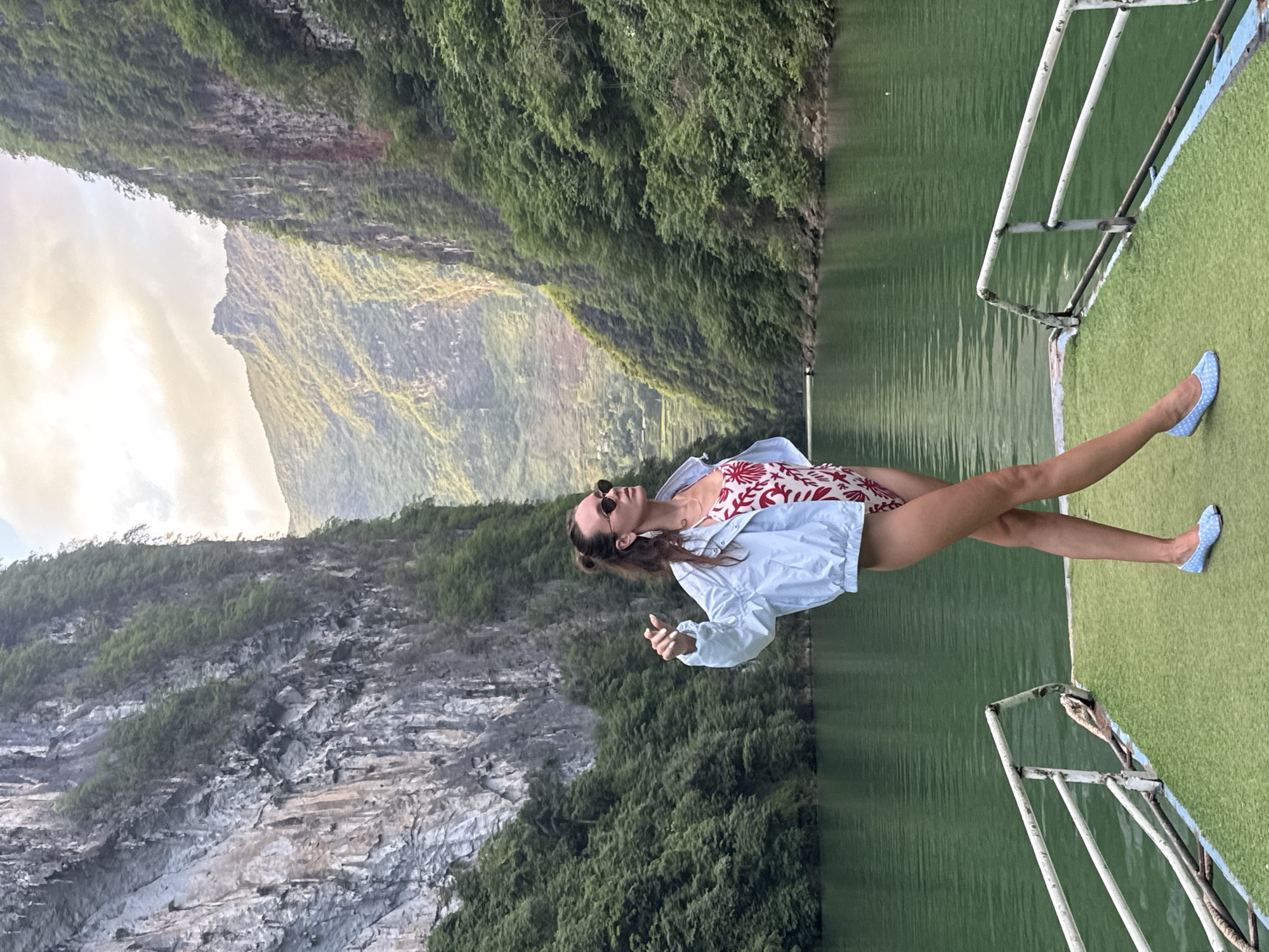 A woman in a red and white swimsuit and light blue shirt stands on a boat, wearing sunglasses. The boat has a green artificial turf surface and white metal railings. She is wearing blue flat shoes with white polka dots. Behind her is a landscape of steep, rocky mountains covered in dense green vegetation. The location is Đồng Văn Commune, Vietnam, as indicated by the landscape. The water is green and calm.