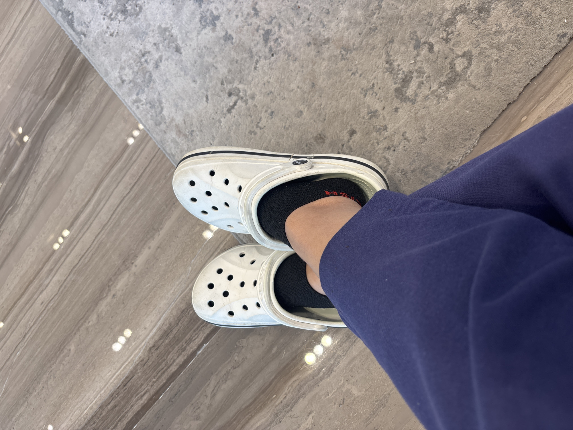 A pair of white Crocs is worn by a person sitting on a floor. The Crocs are white with black trim and black socks are visible underneath. The person is wearing navy blue pants. The floor is a combination of wood-look tile and a textured carpet. The scene is indoors, with lighting reflecting off the tiled floor. The specific location in Dubai, United Arab Emirates is not discernible from the image.