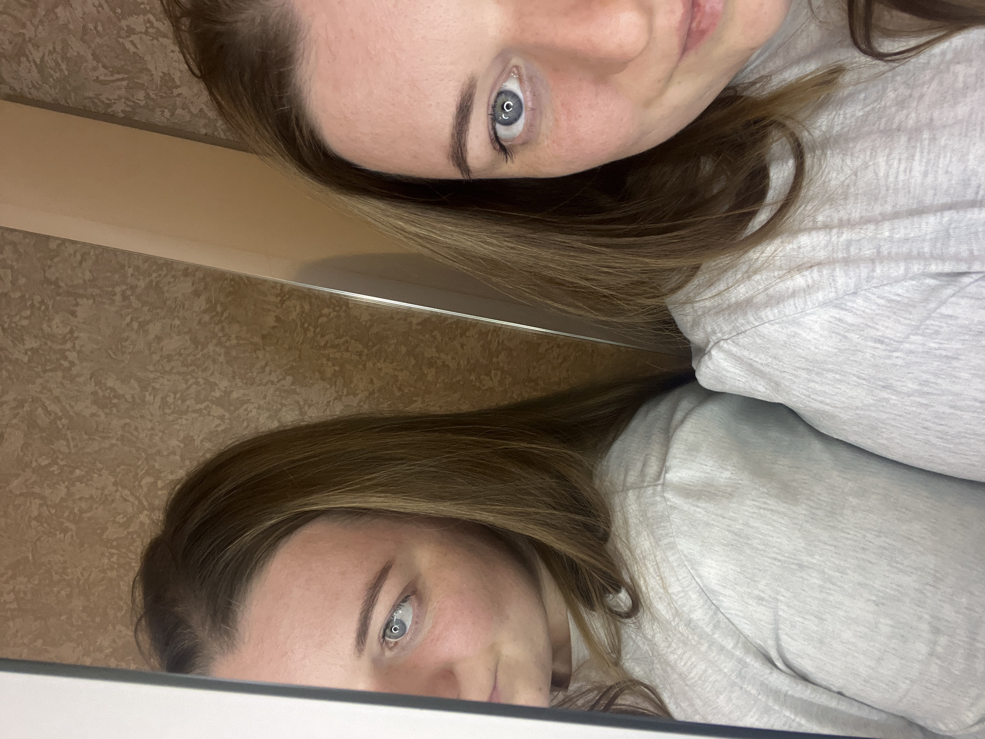 The image shows a woman with fair skin, light brown hair, and blue eyes, reflected in a mirror. She is wearing a light gray shirt. The background behind the mirror shows a textured wall. The location is not discernible from the image.