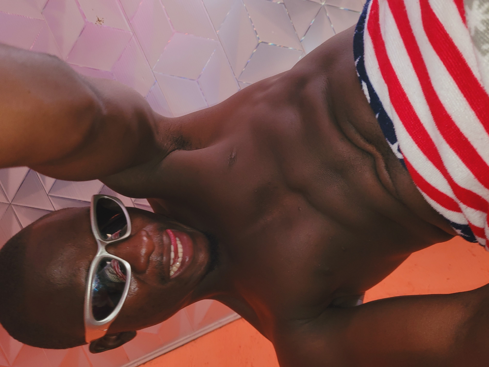 A close-up, rotated portrait captures a dark-skinned individual, appearing to be an adult male, smiling broadly towards the camera. The person is shirtless, showcasing a muscular physique, and their skin appears moist or shiny. They wear silver-framed sunglasses with reflective lenses. A towel or garment, featuring a prominent red, white, and blue striped pattern reminiscent of the American flag, is draped around their lower body on the right side of the frame.
The background features a geometrically patterned wall with reflective tiles, displaying iridescent pink and purple hues under artificial lighting. The lower part of the scene shows a solid orange-peach surface. The individual's wide, open-mouthed smile, revealing teeth and tongue, conveys a joyful and confident emotion. The setting appears to be indoors, possibly a modern space with distinctive tiling, located in Bukuru, Nigeria. No specific activity other than posing is visible. There is no visible text in the image.