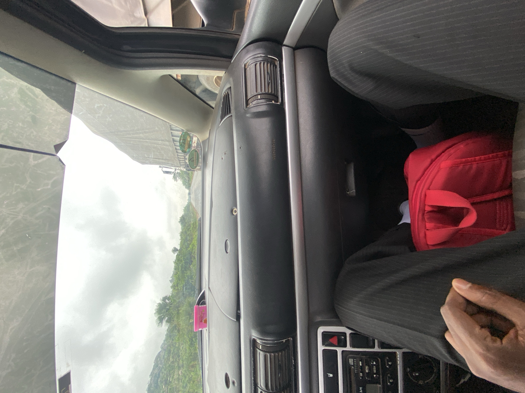The image shows the interior of a car in Assob Hausa, Nigeria, with a view of a green hillside through the windshield. The dashboard is visible, along with air vents and a radio console. A red bag rests on the passenger's side. An individual wearing a pinstripe suit is seated in the car, visible from the waist down. Their hand is resting on their leg. The day is overcast.