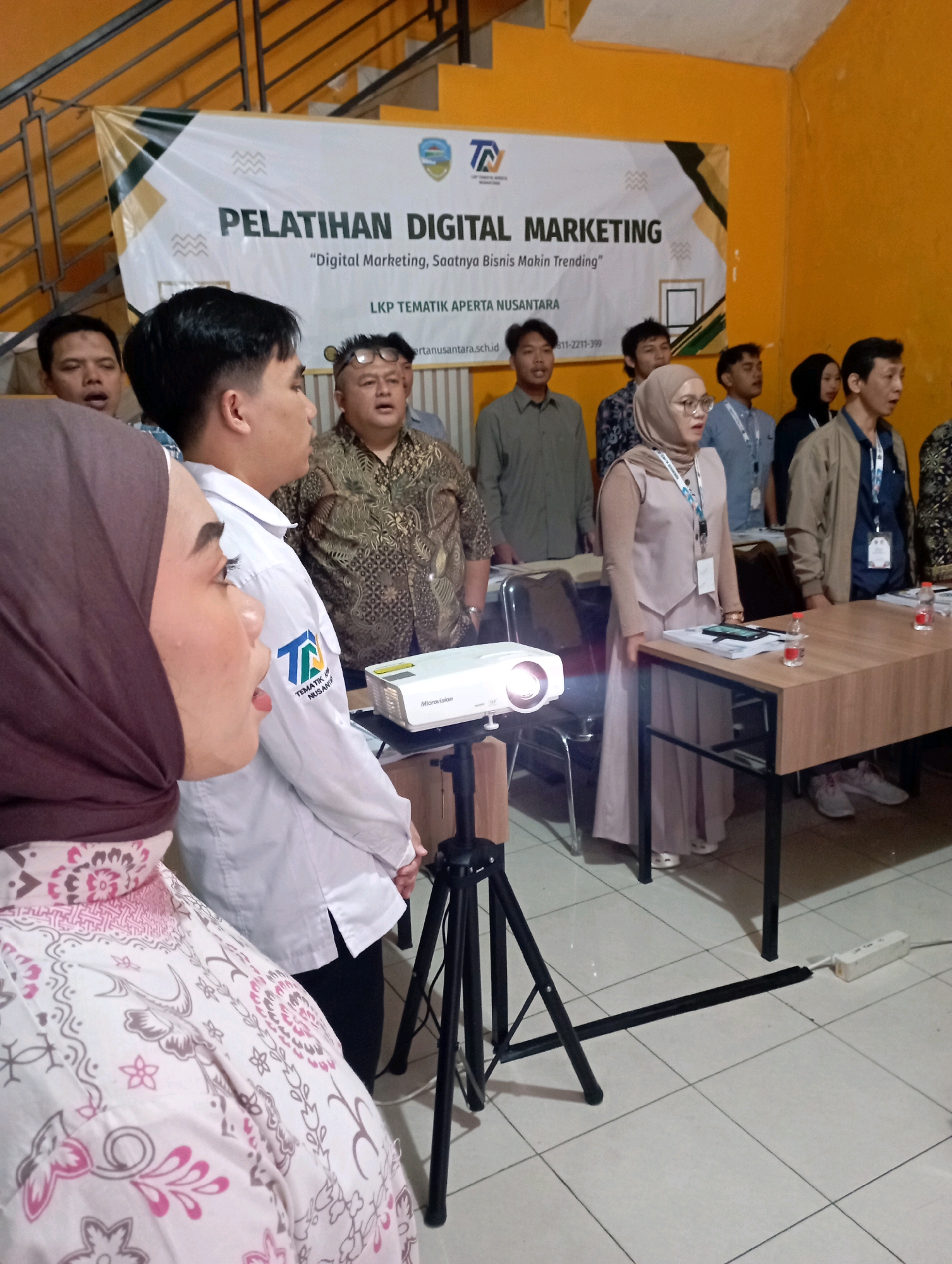 The image depicts an indoor scene, likely a seminar or workshop, where a group of people are gathered. In the foreground, a woman wearing a pink hijab and patterned dress is positioned to the left. Behind her, a man in a white collared shirt stands, also facing the group. A projector is set up on a tripod to the right of the woman. In the background, several other individuals are standing, some in formal attire. A banner is visible on the wall behind the group, with text that appears to be in Indonesian. The environment suggests a professional or educational setting, with tiled flooring and a wall painted in a warm, orange hue. Based on the banner text, the event is related to "PELATIHAN DIGITAL MARKETING" (Digital Marketing Training). The presence of an Indonesian flag emblem on the banner and the language used on the banner suggest the location is in Indonesia. While the image does not explicitly state the city of Tasikmalaya, Indonesia, it's possible to infer this location if external information links this event or organization to Tasikmalaya.
