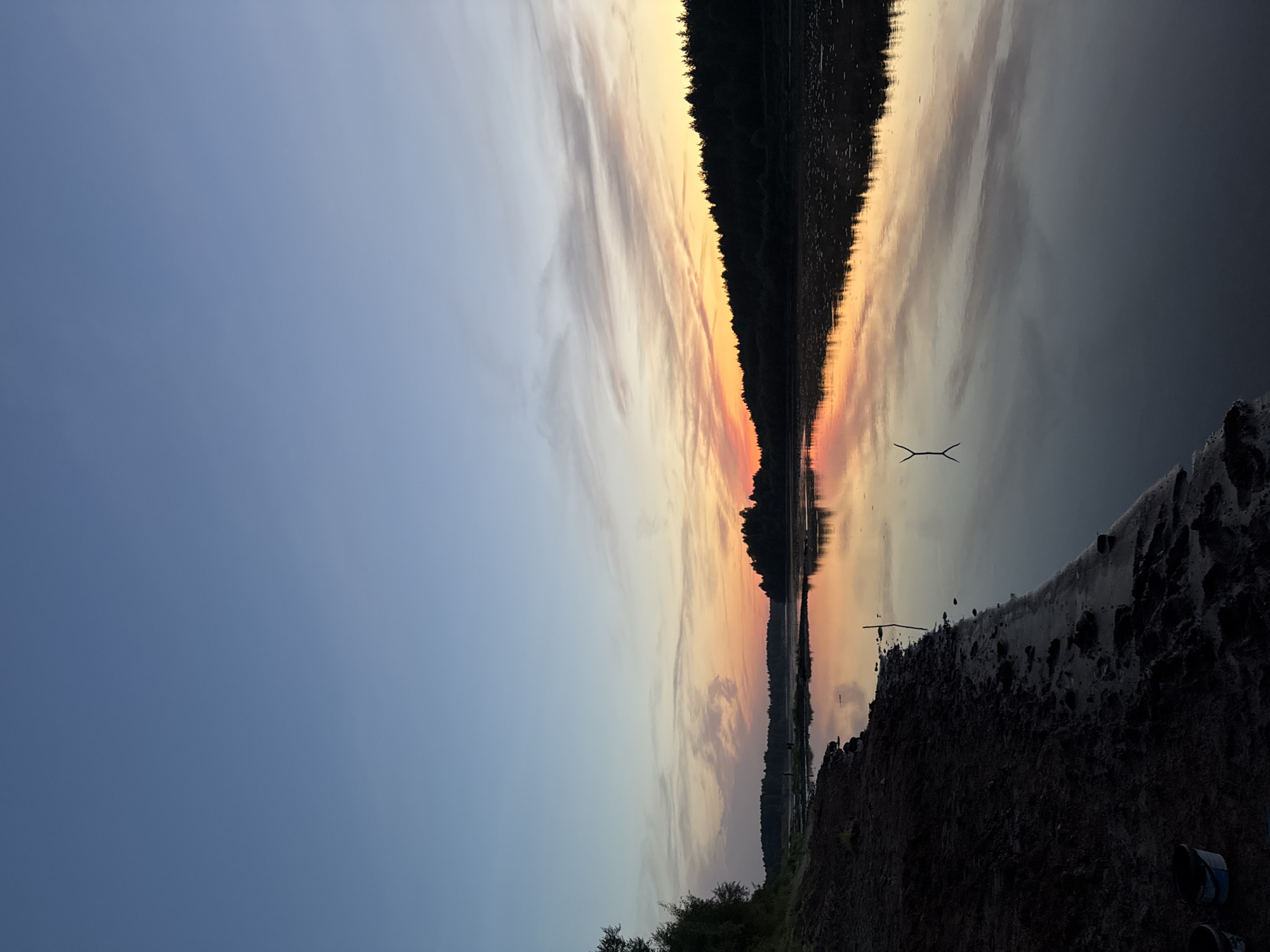 The image depicts a serene natural landscape at dusk, likely near the city of Syktyvkar, Russia, given the context. The foreground features a muddy shoreline with some debris and two white buckets visible at the bottom left. To the right, a calm body of water reflects the sky, which is transitioning from a clear blue overhead to hues of orange, pink, and yellow near the horizon, indicating sunset. Wispy clouds are scattered across the sky, further enhancing the colorful display.
A dense line of dark trees silhouettes the opposite bank of the river, creating a stark contrast against the vibrant sky. In the distance, several indistinct figures can be seen on the water or near the shoreline, suggesting human presence. A single stick or branch stands vertically in the water, its reflection mirrored below. The overall atmosphere is tranquil and suggestive of a peaceful evening by the water.