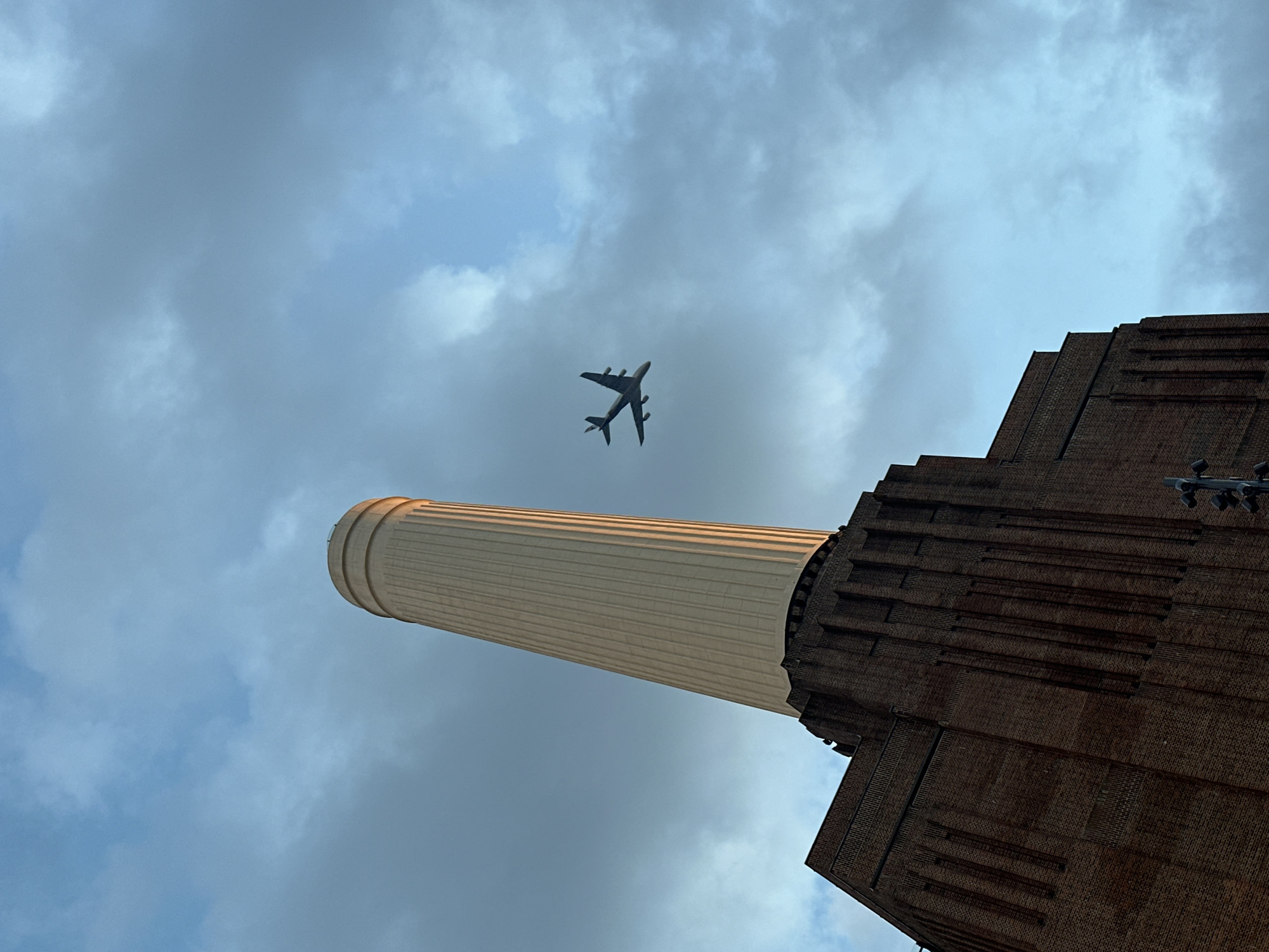 The image shows a low-angle view of the Battersea Power Station in London, United Kingdom, under a cloudy sky. A large passenger airplane is visible in the sky between one of the chimneys and the building structure. The power station's brick facade is prominent, along with one of its iconic white chimneys. The sky consists of a mix of blue patches and grey clouds. There are no people visible in the image.