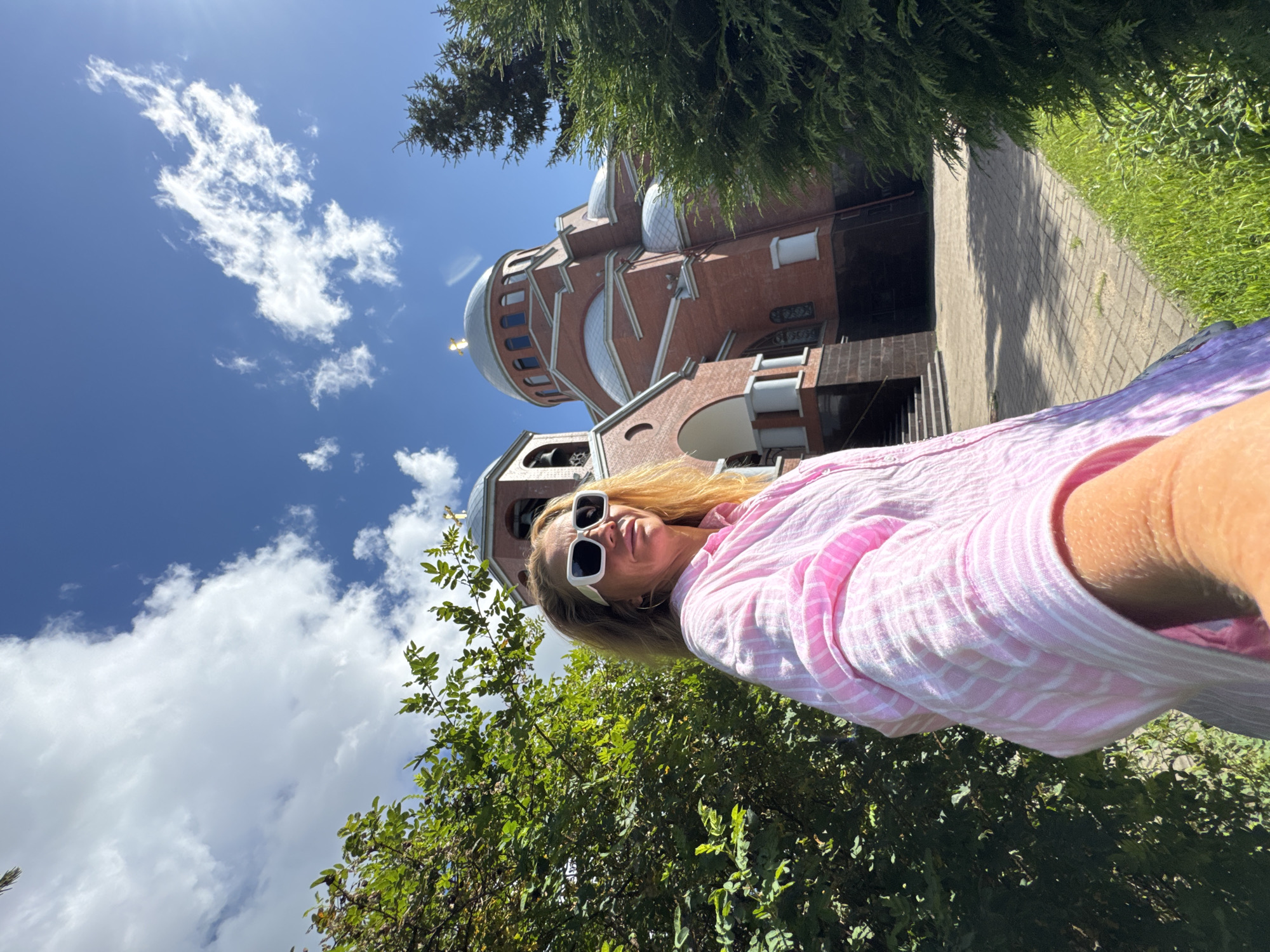 In Saint Petersburg, Russia, on a sunny day with scattered clouds, a woman with blonde hair and white sunglasses is taking a selfie outdoors. She is wearing a pink and white striped shirt. Behind her, a red brick church with domes and a bell tower is visible. Lush green trees and bushes surround the church and the woman. A paved walkway leads to the church entrance.