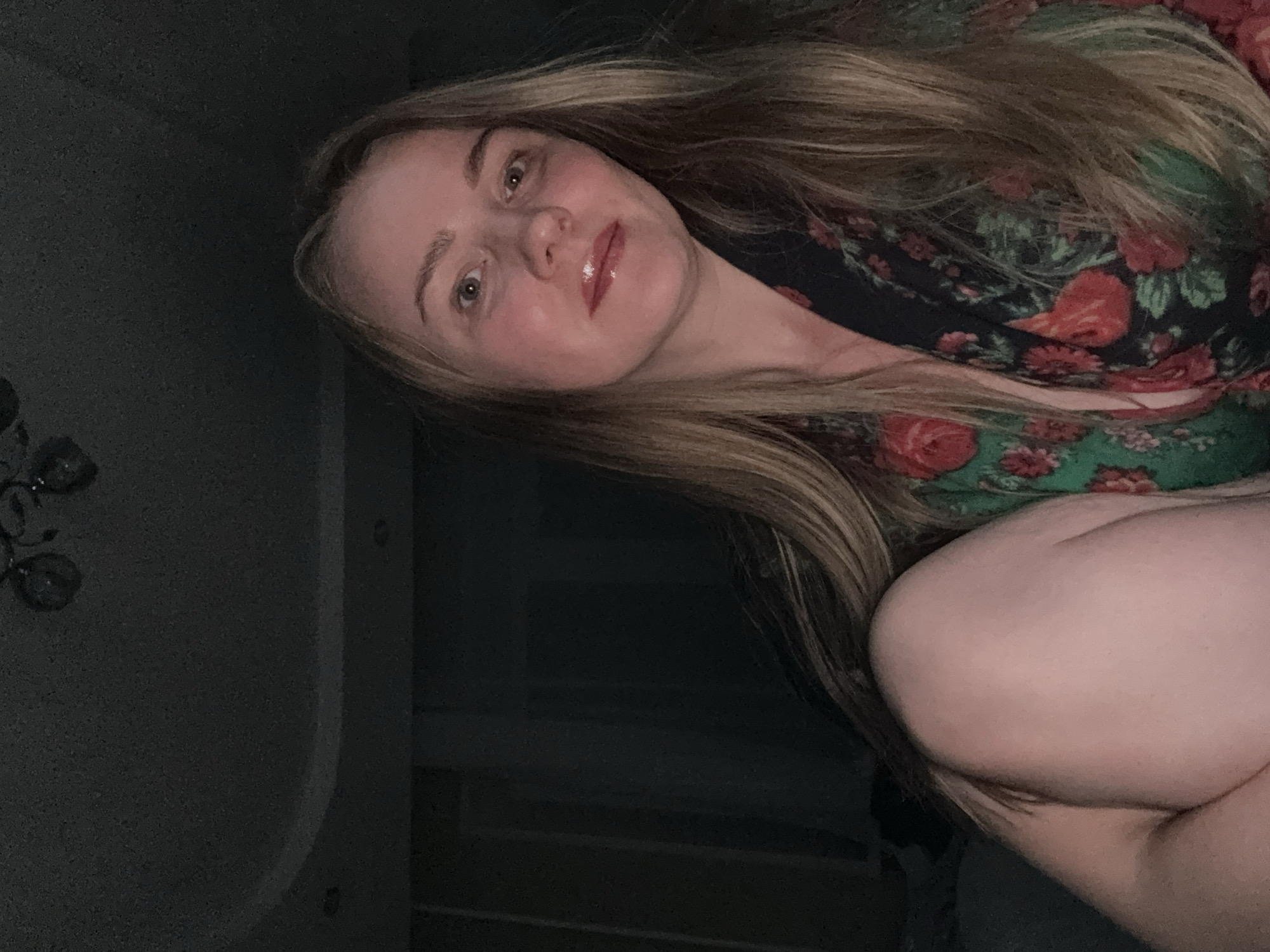 The image features a young to middle-aged woman posing for a close-up photograph, rotated 90 degrees clockwise. She has long, flowing blonde or light brown hair, which frames her face. Her light-colored eyes, possibly green or hazel, look directly at the viewer with a soft, relaxed expression and a slight smile. Her lips are adorned with a light pink or clear lip gloss.
She is wearing a garment, likely a top or dress, characterized by a vibrant floral pattern. The fabric is dark, possibly black or deep green, with prominent red flowers and green leaves scattered across it. Her posture suggests she is either sitting or reclining comfortably. A part of her arm or leg is visible in the lower right, out of focus.
The setting appears to be indoors, dimly lit, suggesting evening or night. The background is mostly dark and indistinct, with a decorative light fixture or chandelier faintly visible in the upper left corner. The overall mood conveyed by the soft lighting and her gentle expression is one of calm and comfort. There is no visible text in the image.