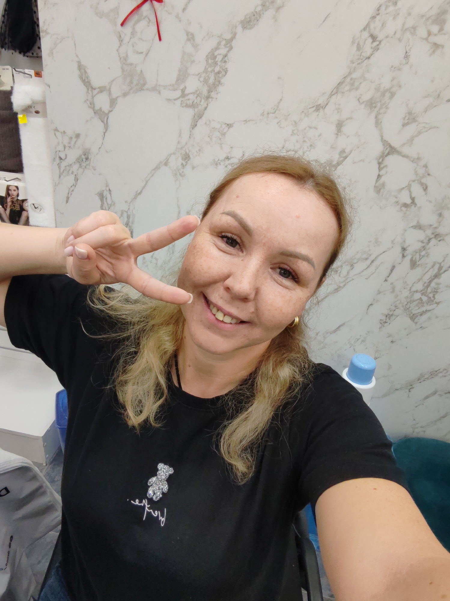 A woman, likely in Chaikovsky, Russia, is captured in a cheerful selfie. She smiles broadly at the camera while making a peace sign with her right hand. Her blonde hair is loosely styled, and her face is adorned with freckles. She wears a black short-sleeved t-shirt featuring a sparkling silver teddy bear design and stylized cursive text underneath, which appears to read "love." Her nails have a neat French manicure, and she wears a gold hoop earring.
The background consists primarily of a light-colored wall with a distinctive gray marble-like pattern. To the left, various items are visible, including white fluffy material, product packaging with images of a woman, and partially visible text, notably the word "BAD" on one package. A small red ribbon is pinned to the wall in the upper left corner. On the right side of the frame, behind the woman's shoulder, a white and blue bottle or dispenser can be seen, along with a glimpse of a teal piece of furniture. The lighting suggests an indoor setting.